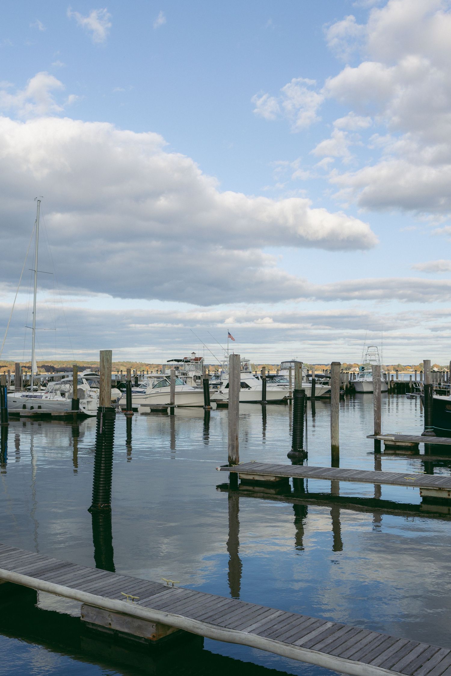 Calm marina with wooden docks, moored sailboats and yachts reflecting in still water under a partly cloudy blue sky.