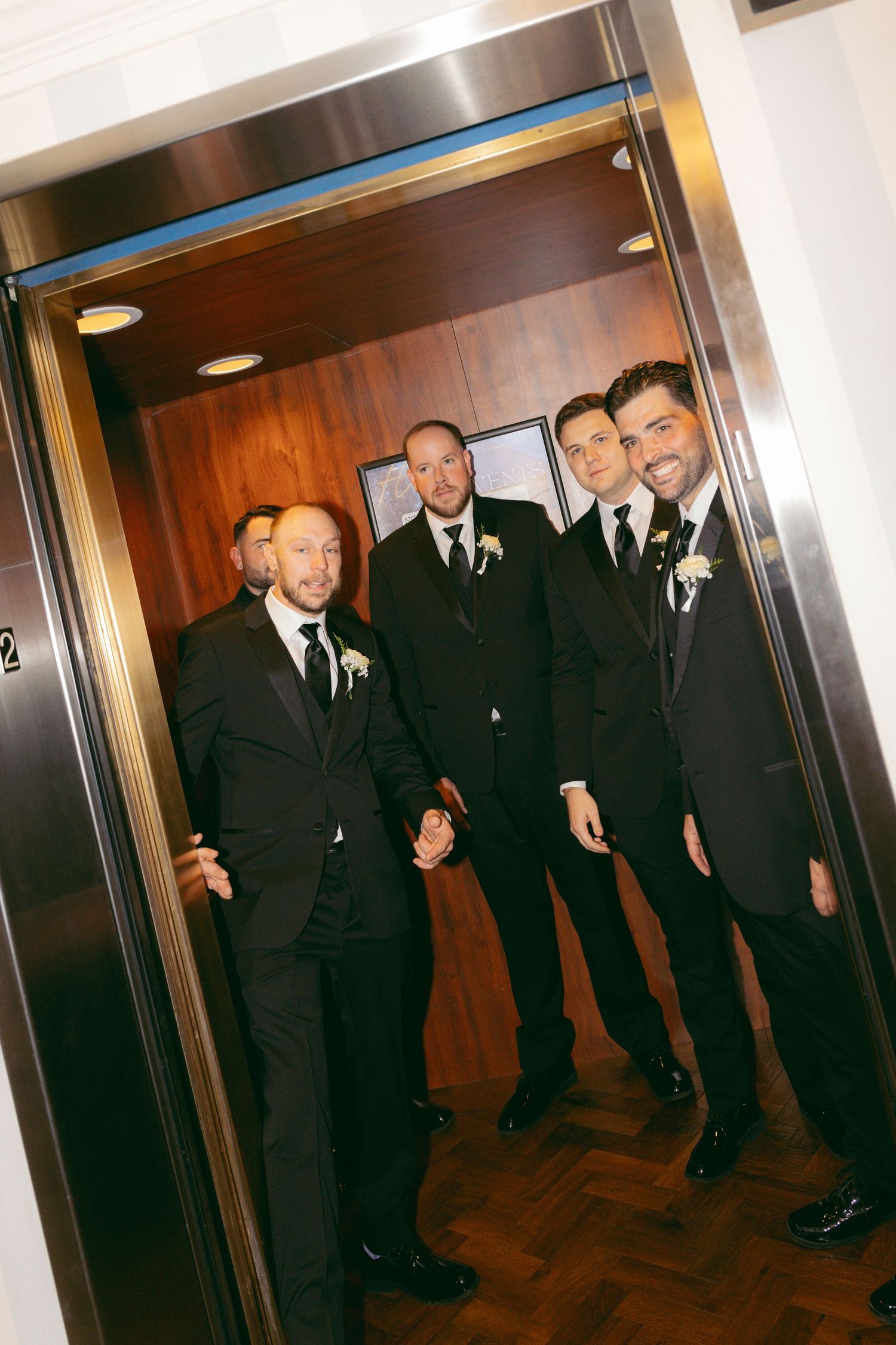 Four groomsmen in black suits with boutonnieres posing inside an elevator at a wedding venue.
