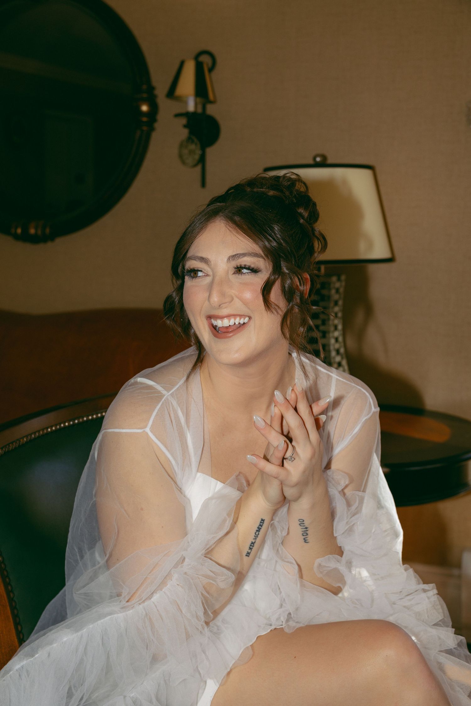 Smiling bride in white sheer robe with updo hairstyle and tattoos, sitting in elegant hotel room before wedding.