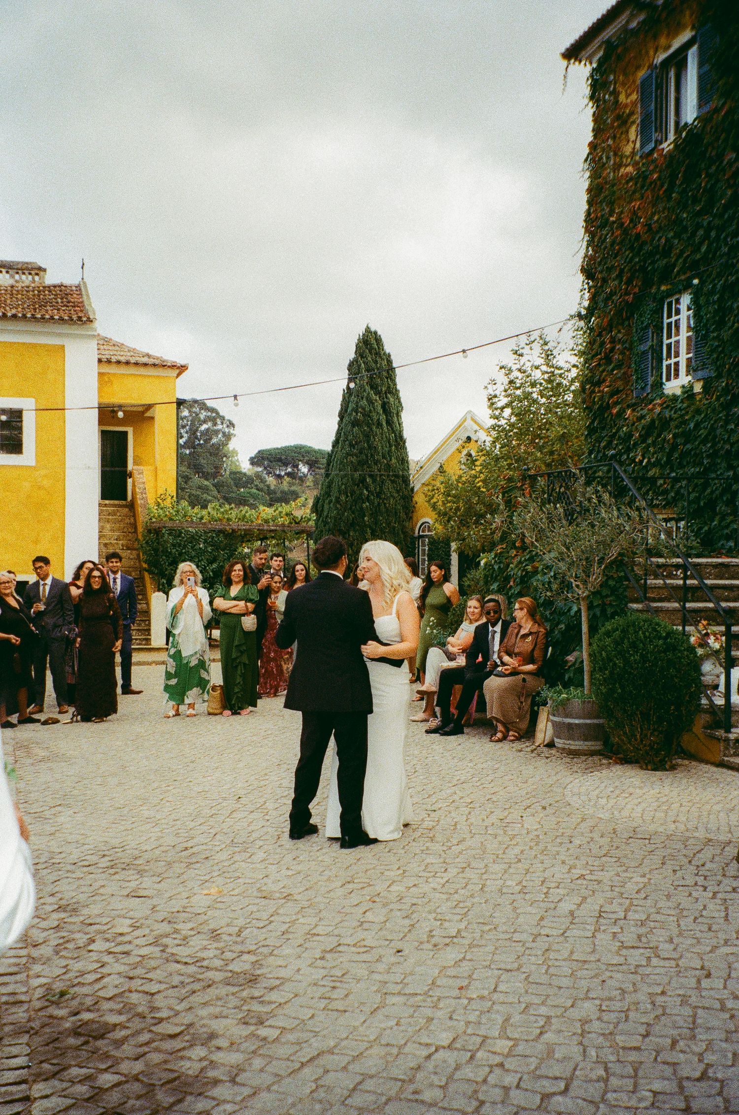 Bride and groom share first dance outdoors at rustic European venue with ivy-covered walls and guests watching.
