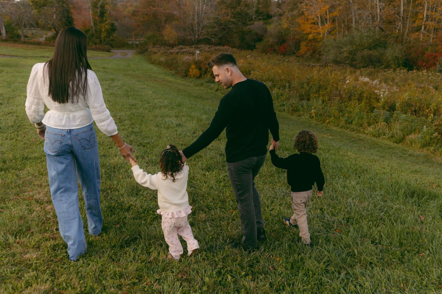 Family of four holding hands walking through a green meadow with autumn trees in the background.