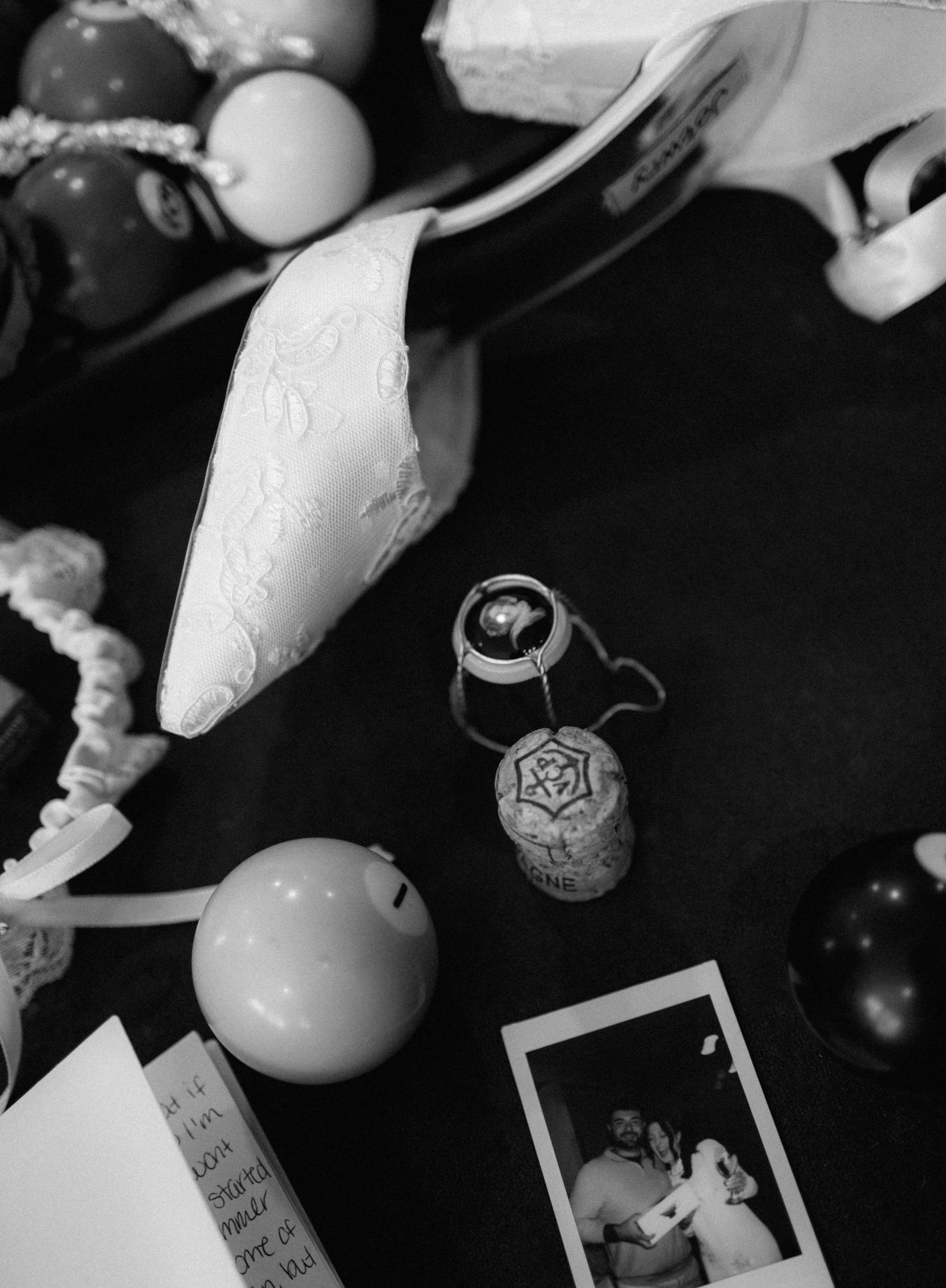 Black and white flat lay of wedding shoe, champagne cork, Polaroid photo, and decorative balls on dark surface.