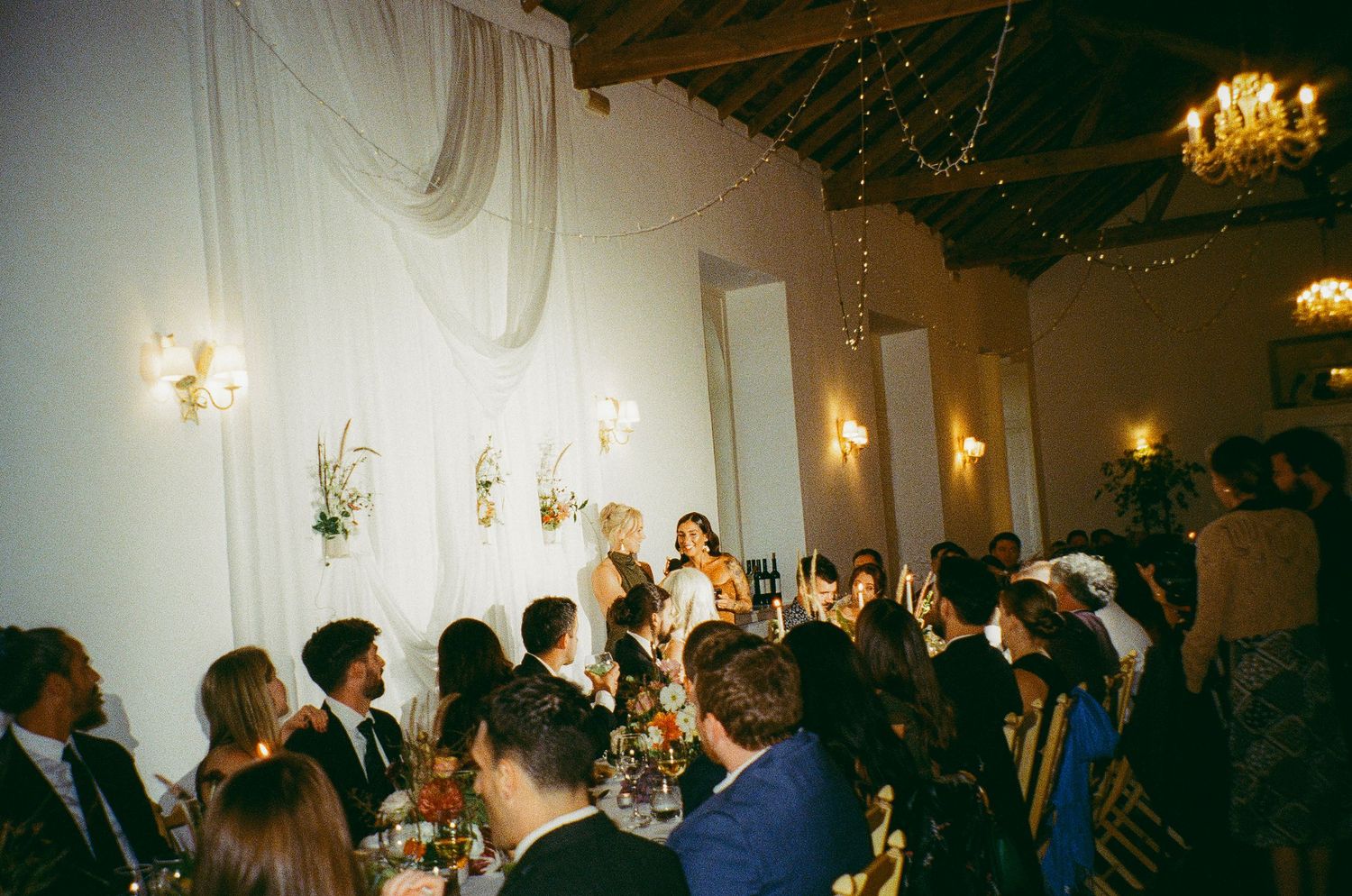 Elegant wedding reception dinner in a rustic venue with warm lighting, guests raising glasses during a toast.