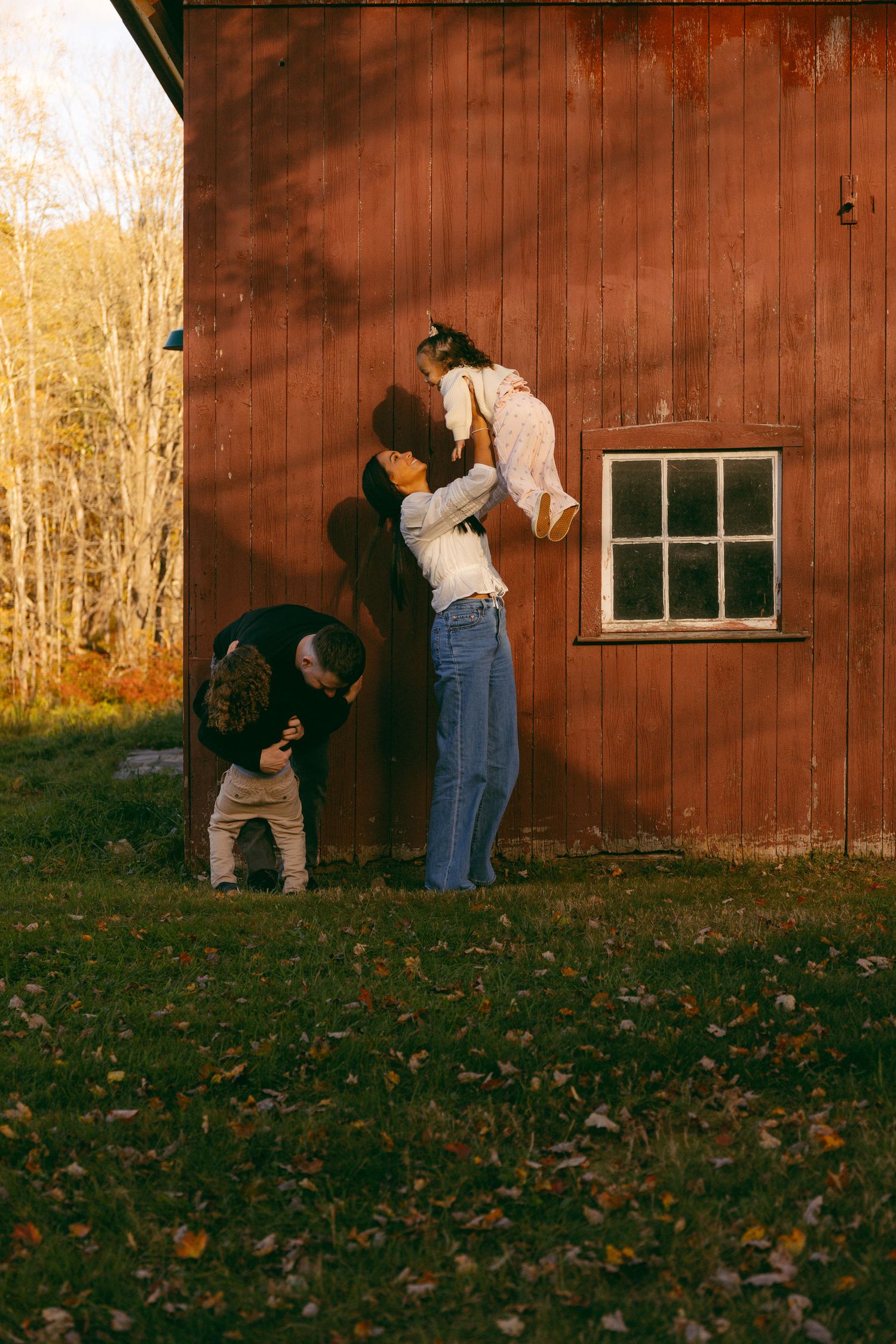 Family playing outdoors by a red barn in autumn, mother lifting baby while father bends down near fallen leaves.