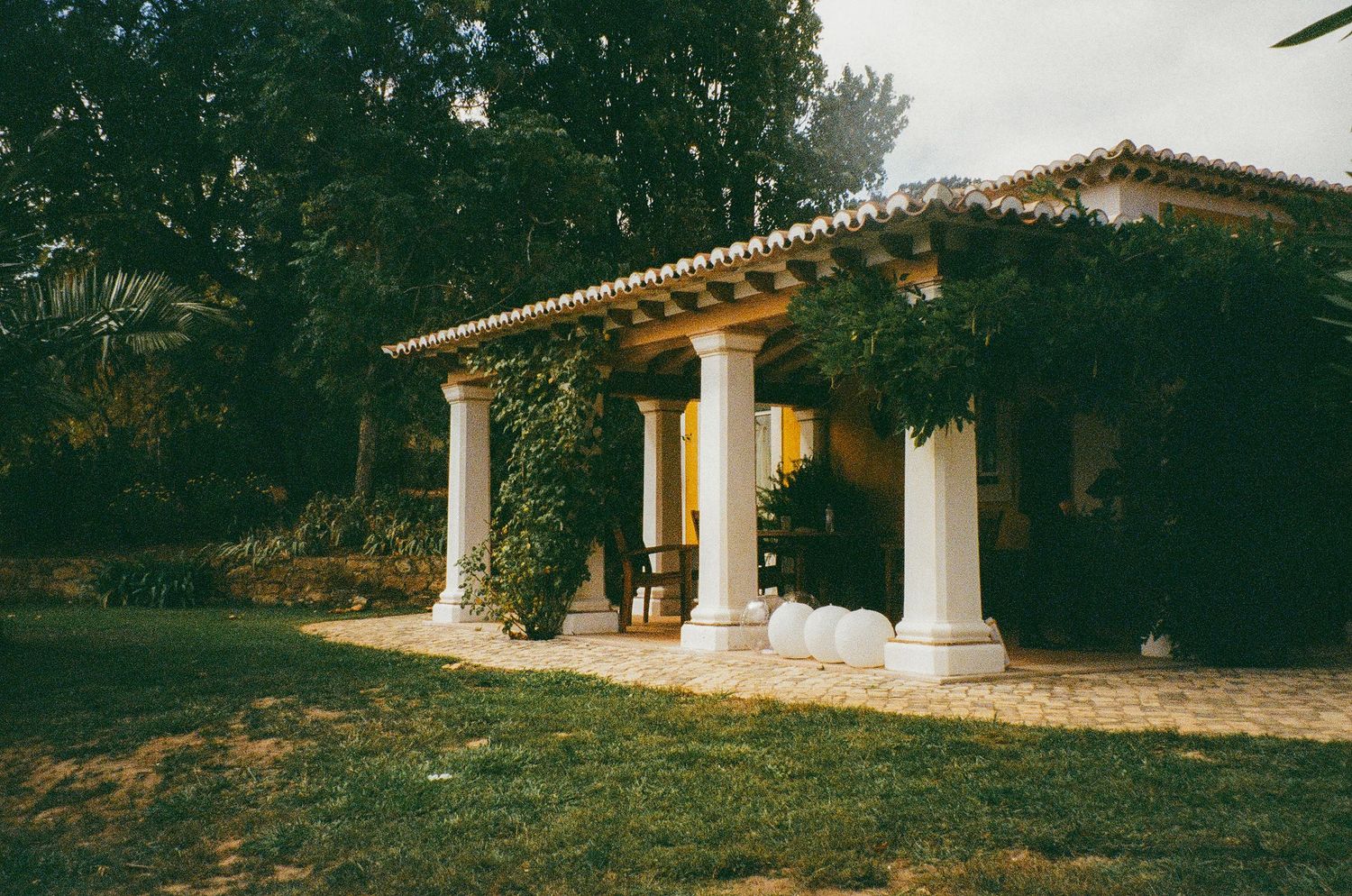 A charming villa with white columns and ivy-covered pergola surrounded by lush green trees and manicured lawn.