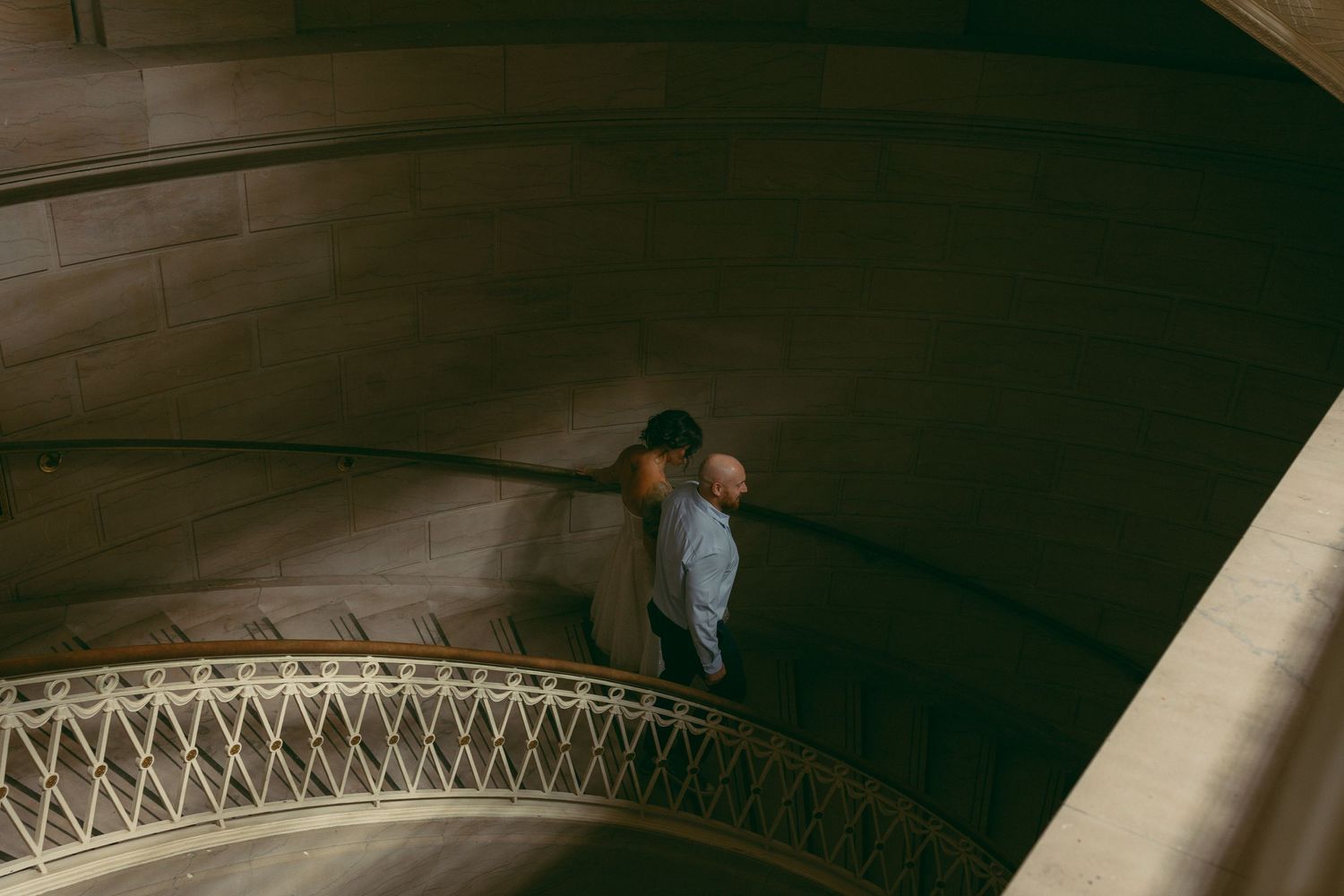 A person stands on an ornate spiral staircase with decorative iron railings in a grand architectural interior.