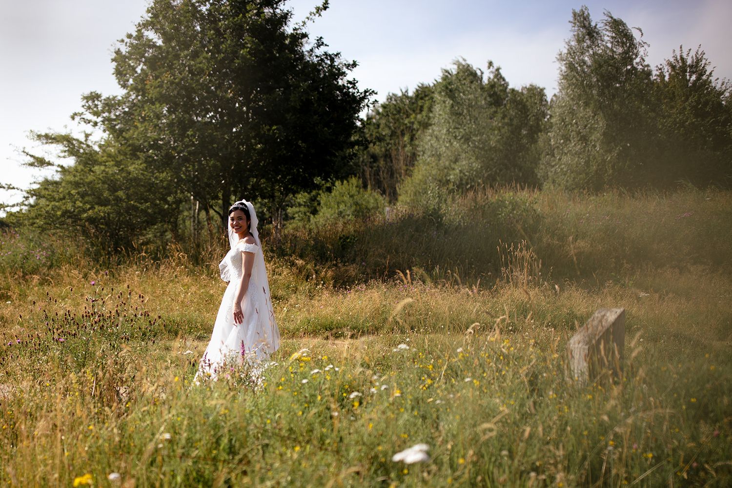 Bride in white wedding dress and veil standing in a lush green meadow with wildflowers and trees in background.