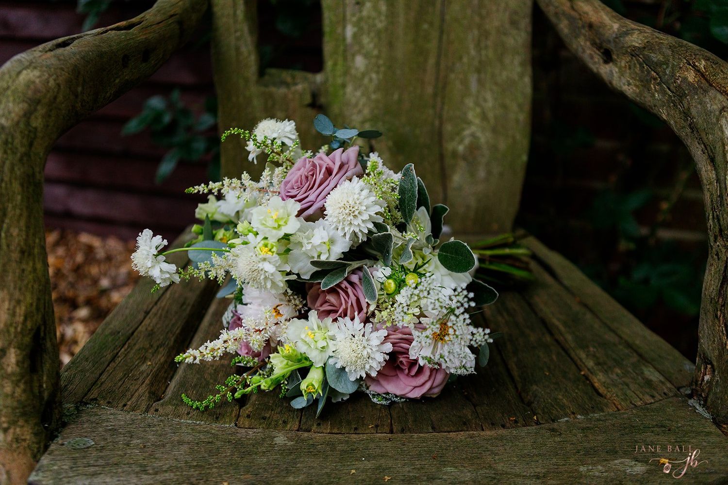 A rustic wedding bouquet with white and pink flowers rests on a weathered wooden bench in a garden setting.