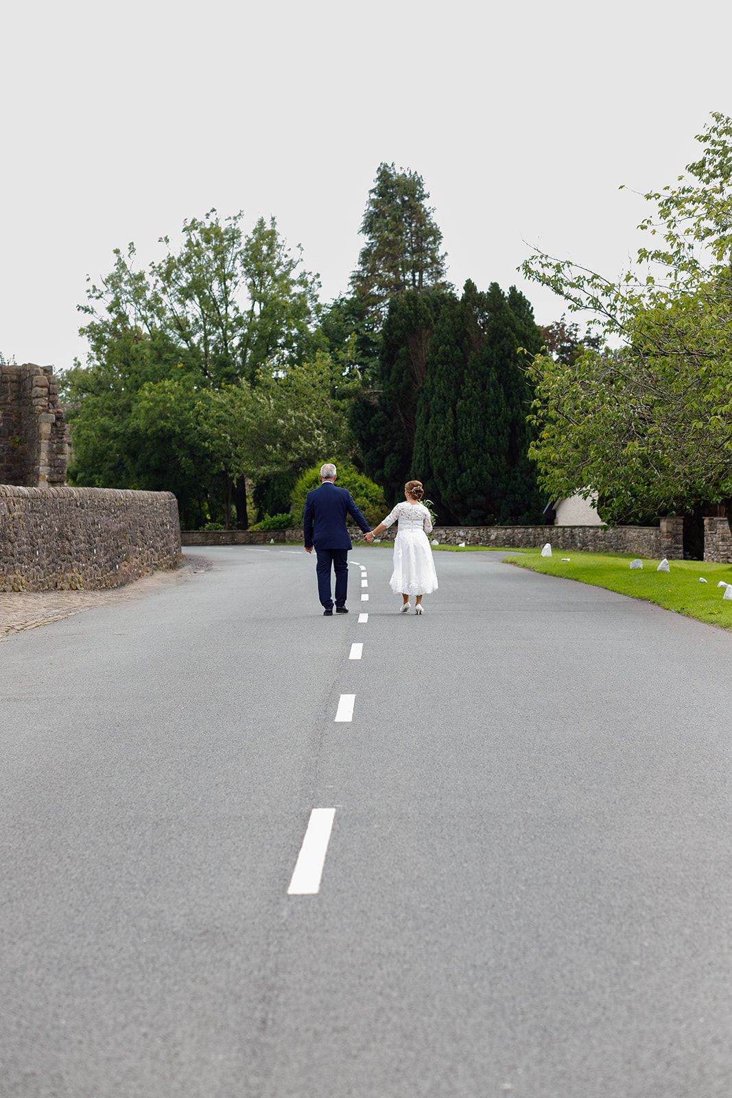 Two people walk together on a long paved path bordered by stone walls and green trees on a cloudy day.