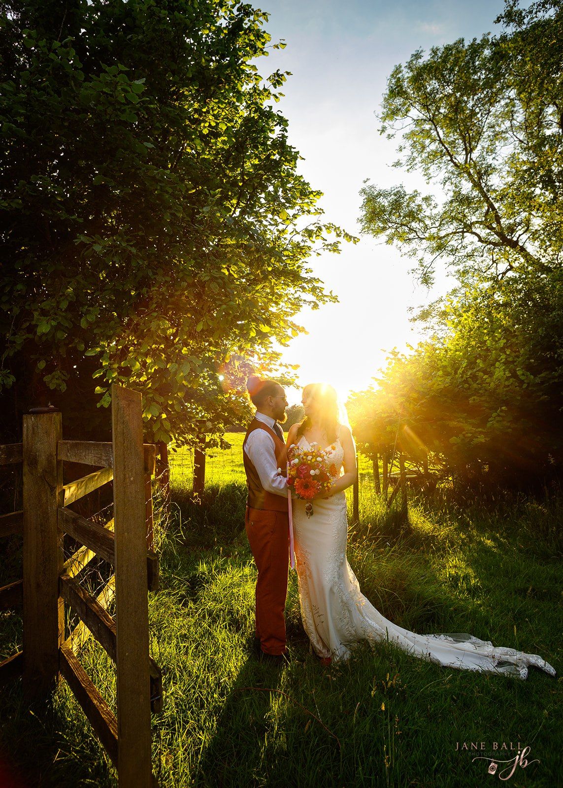 Bride and groom kissing at sunset in lush green countryside, golden light glowing through trees on their wedding day.