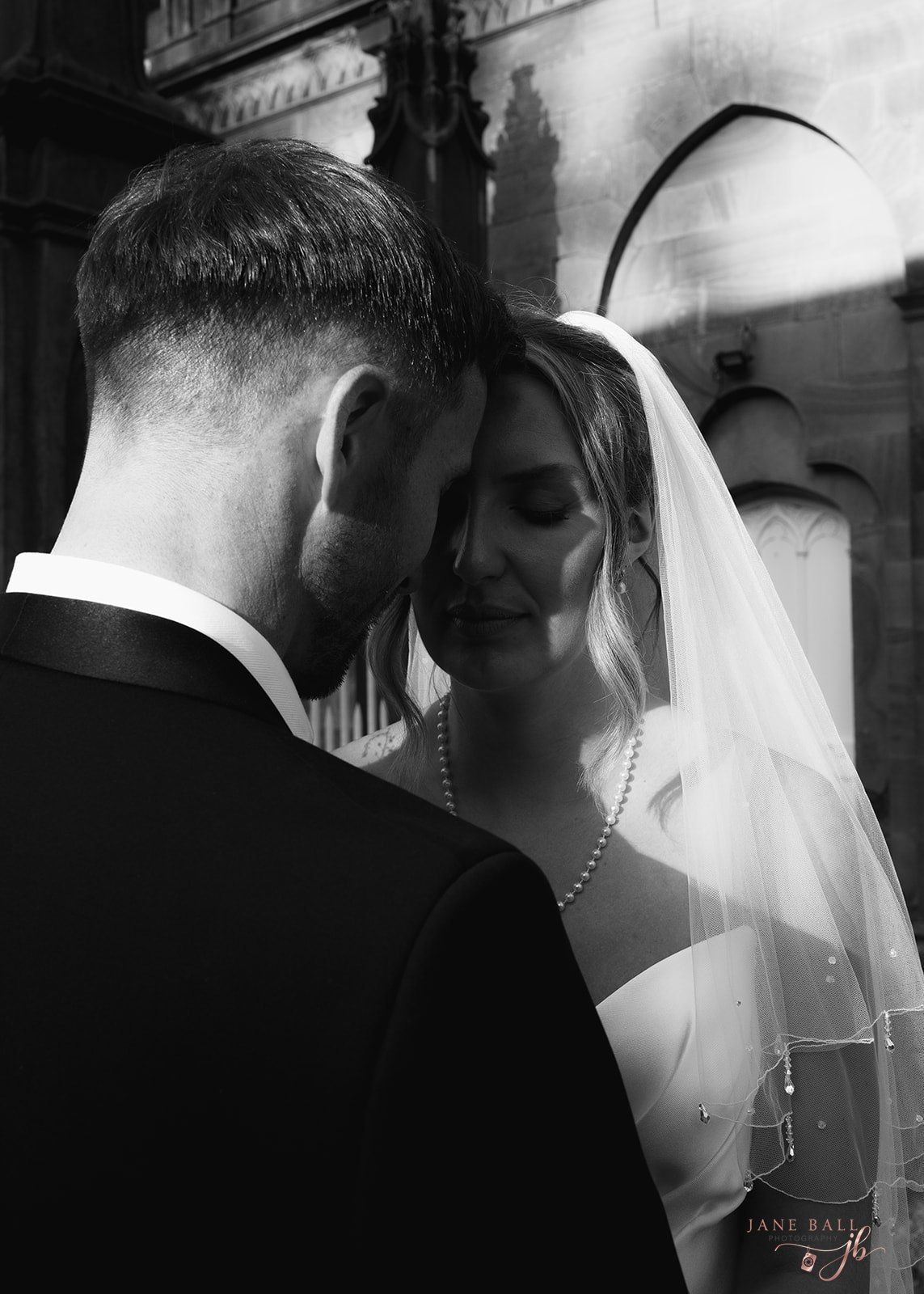 Black and white intimate wedding photo of bride in veil and groom in tuxedo sharing a tender moment in church setting.