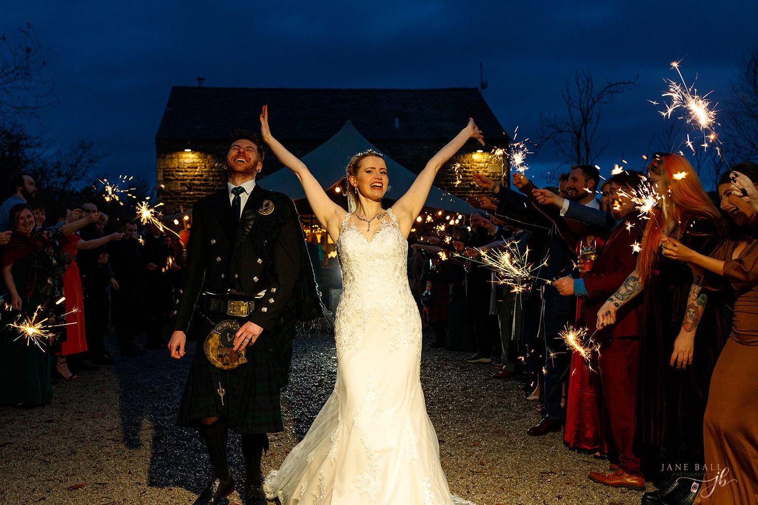 Bride in a white gown raises her arms beside a groom in a kilt as wedding guests hold sparklers at an evening outdoor celebration.