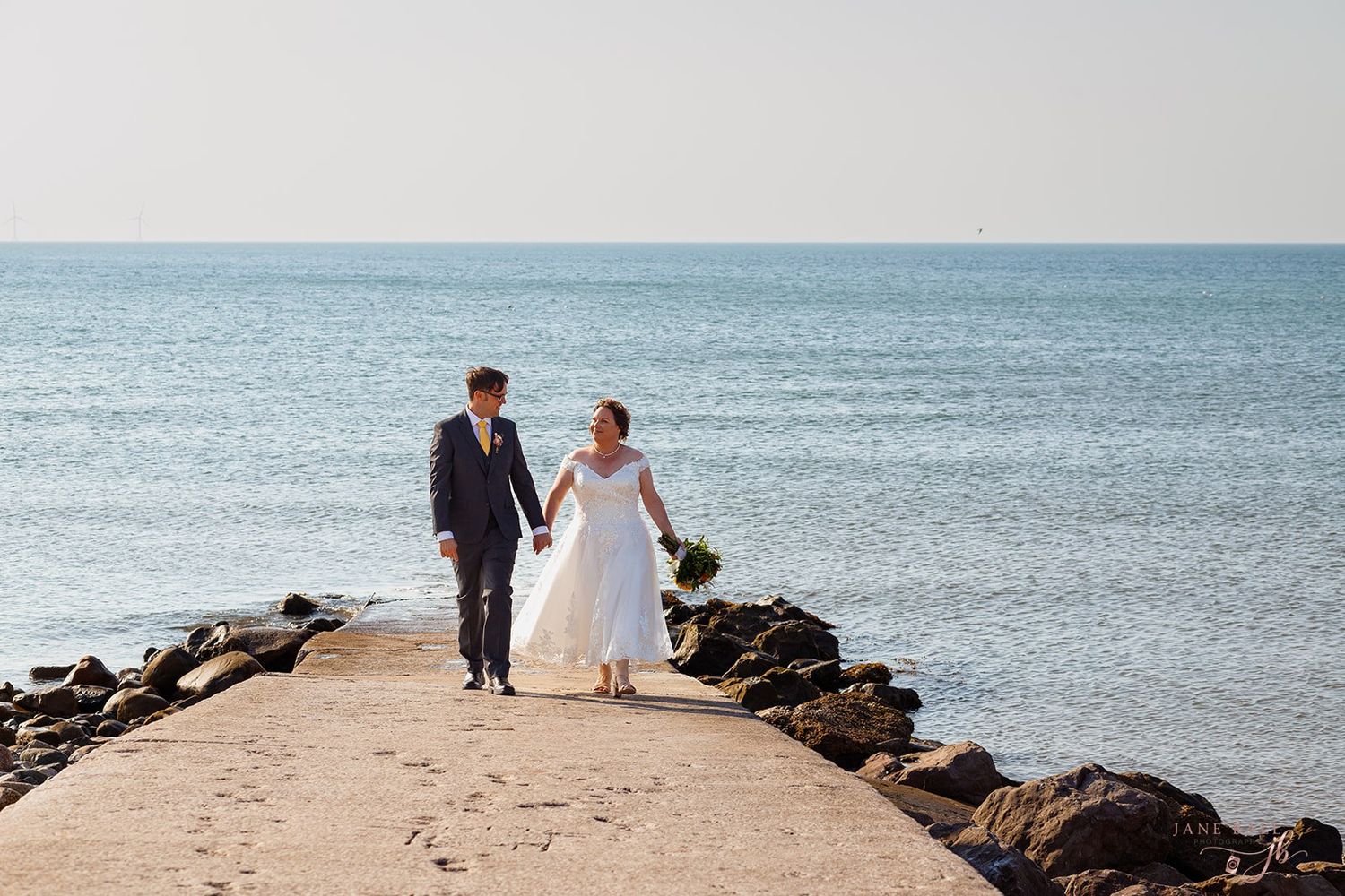 Couple in wedding attire walking along a rocky beach shore with ocean waves in the background.