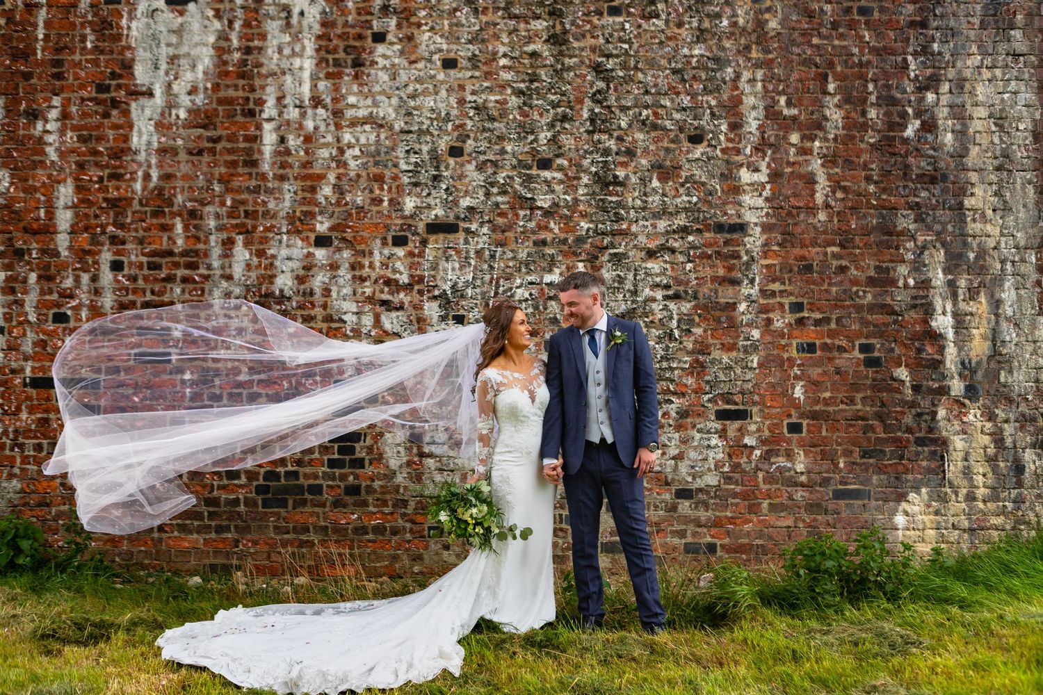 Wedding couple posing against rustic brick wall with flowing veil in the wind.