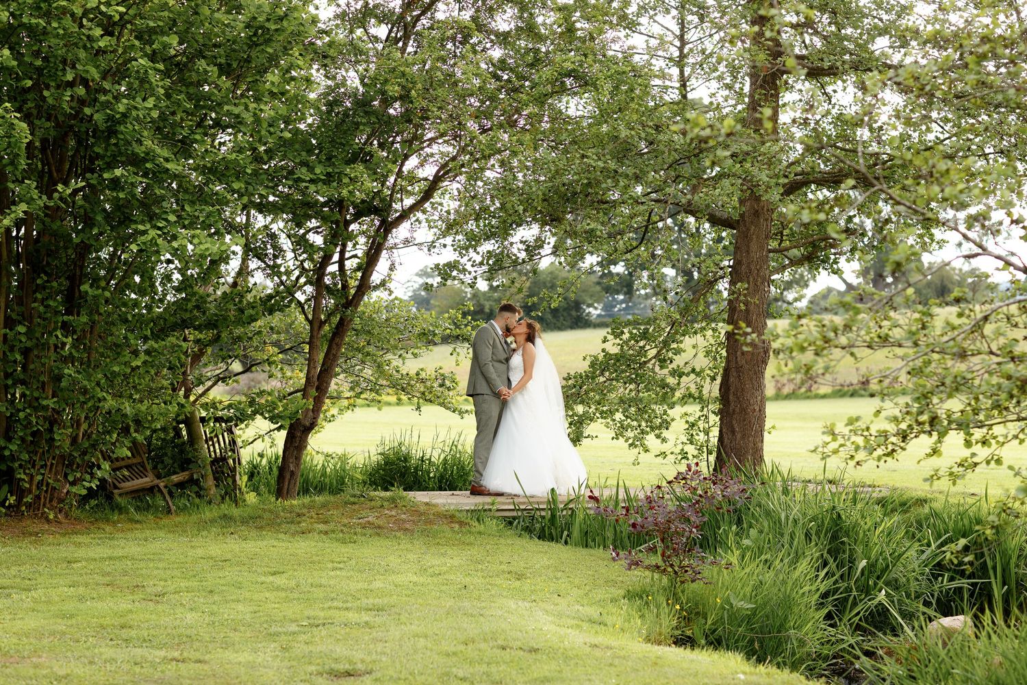 The couple embraces outdoors surrounded by lush greenery and trees in a romantic garden setting.