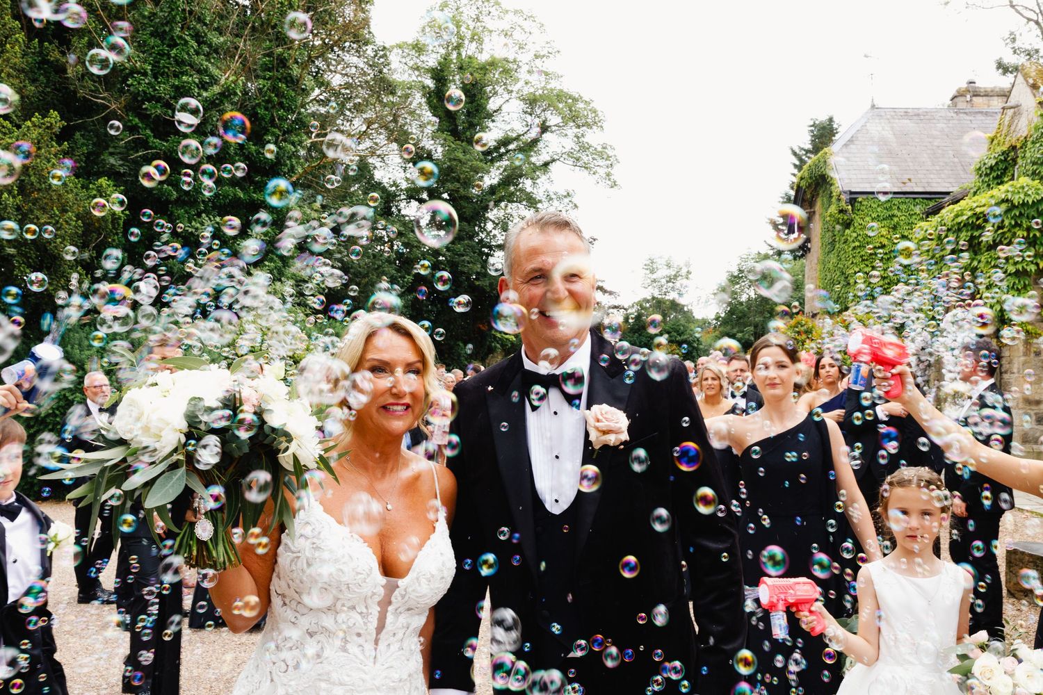Newlyweds walk through a shower of bubbles surrounded by guests during an outdoor wedding celebration.