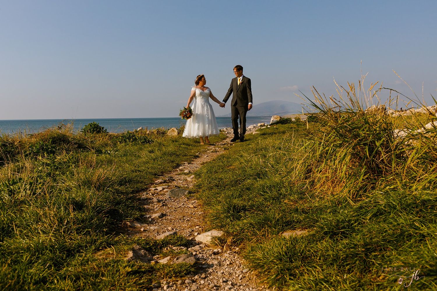 A couple in wedding attire walks along a grassy coastal path overlooking the ocean on a sunny day.