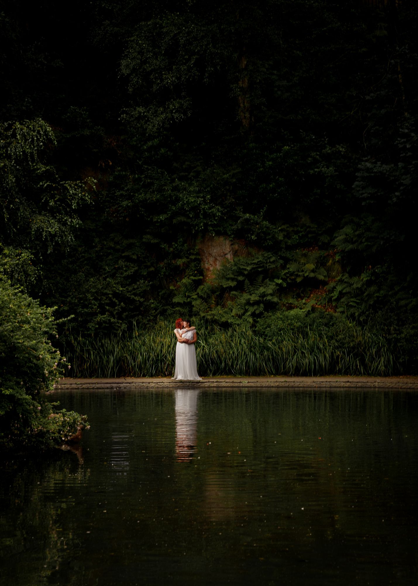 A romantic wedding couple stands in a serene lake surrounded by lush greenery and trees in moody lighting.