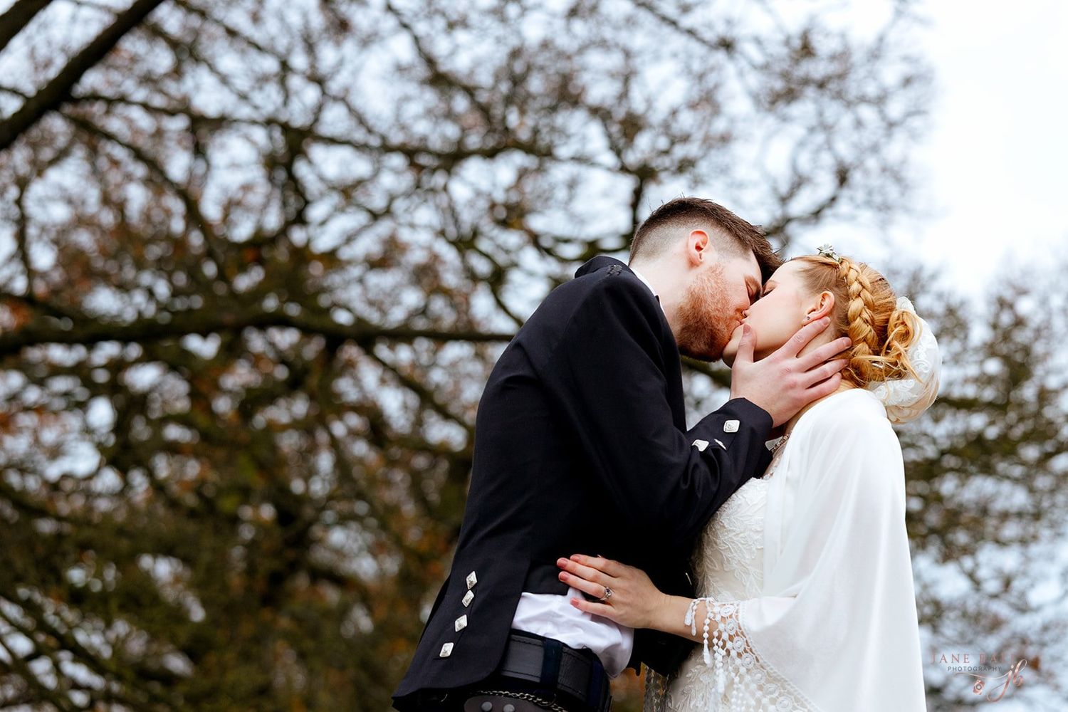 A romantic wedding couple embraces outdoors in a winter scene with bare trees in the background.