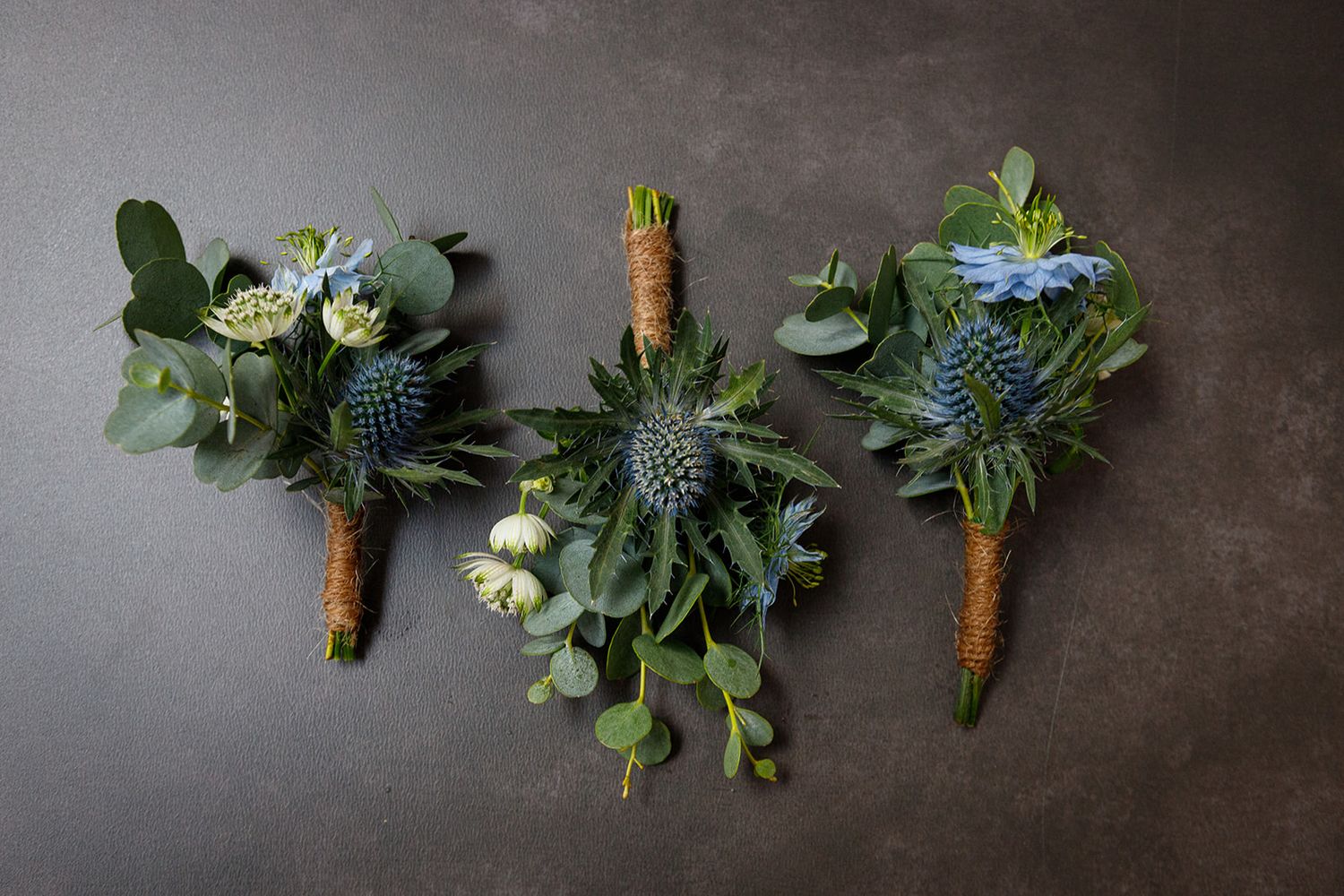 Three rustic wedding boutonnieres with blue thistle flowers, white blooms and greenery wrapped in twine on dark surface.