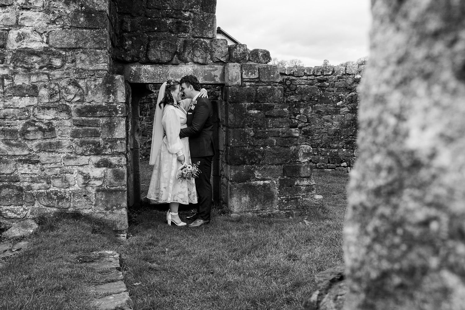 Black and white wedding photo of couple kissing among ancient stone ruins, bride in lace dress, romantic outdoor ceremony.