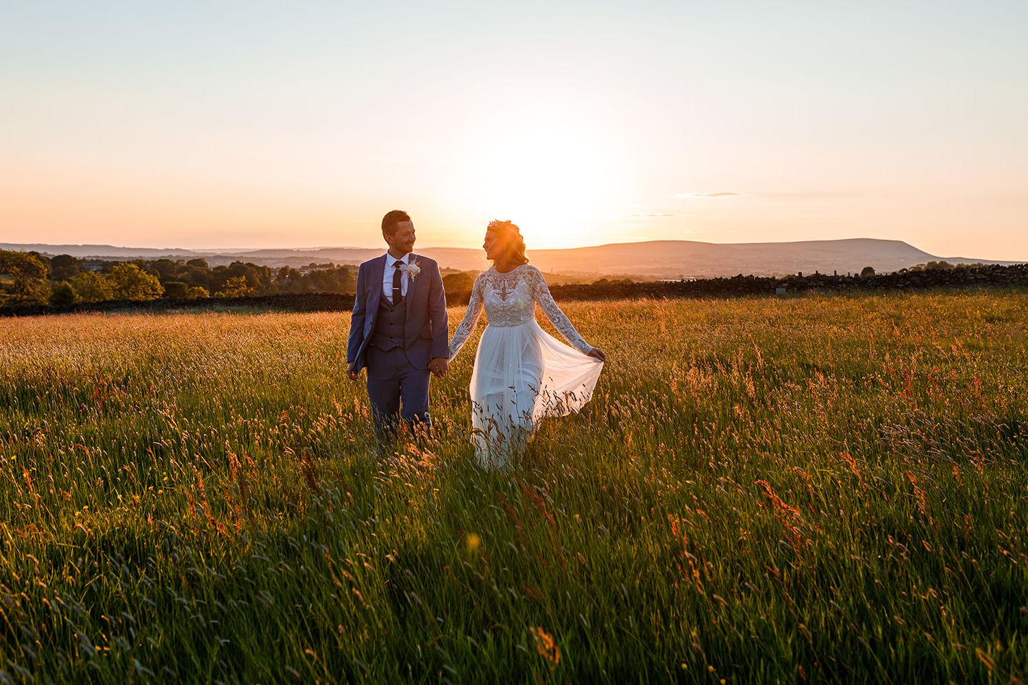 Two figures walk hand in hand through a golden field at sunset with mountains in the background.