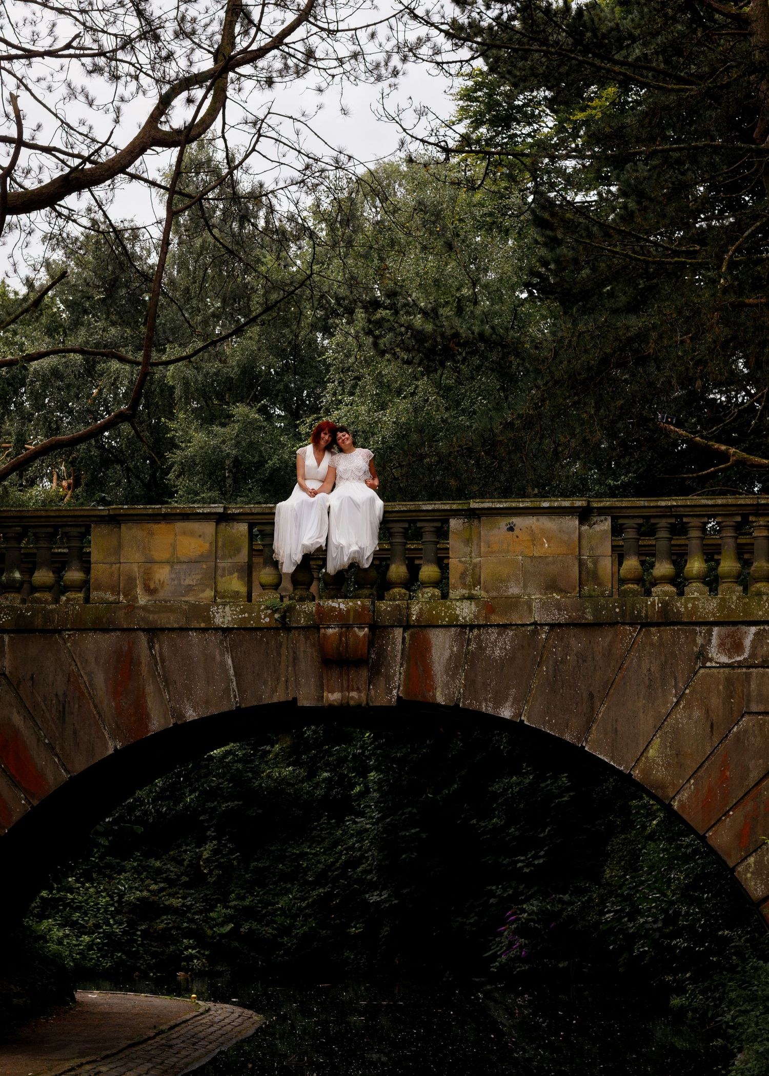 Two brides in white dresses sit together on a stone bridge arch surrounded by lush green trees.