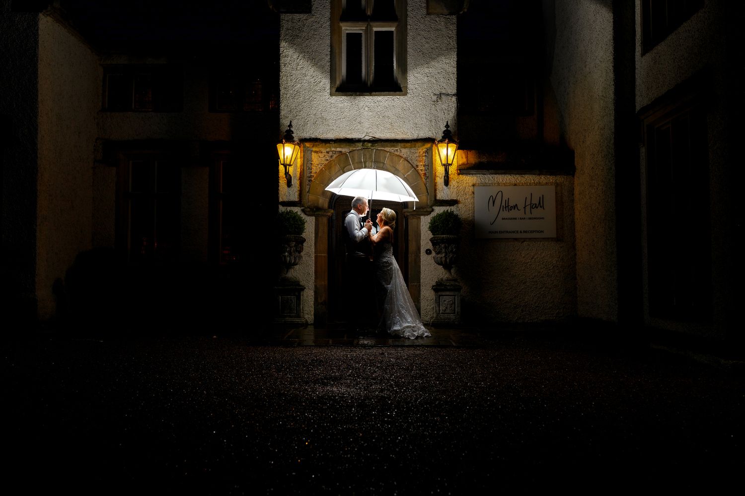 A romantic wedding photo of a couple under a white umbrella at night with glowing lanterns in the background.