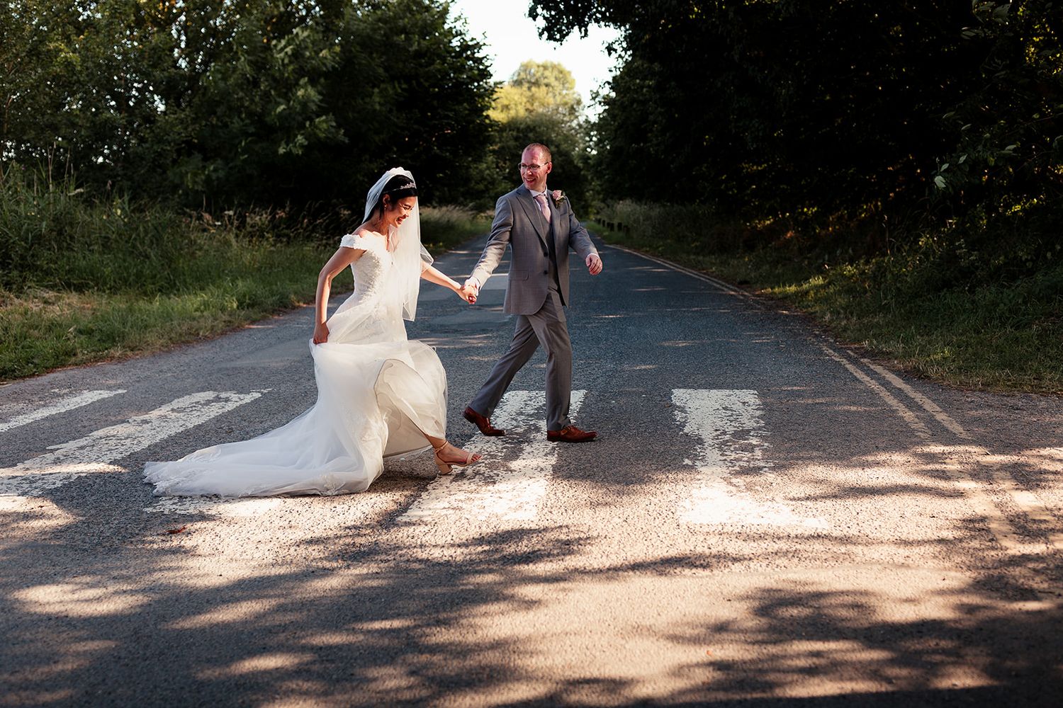 Bride in a white wedding gown holding hands with a groom in a gray suit as they walk across a sunlit tree-lined road.