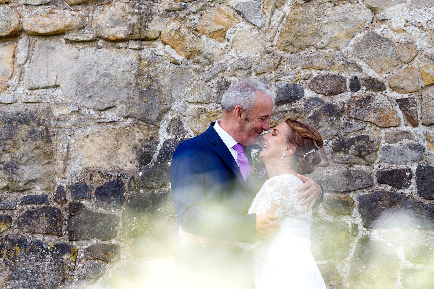 A romantic wedding couple shares an intimate moment against a rustic stone wall with soft glowing light effects.