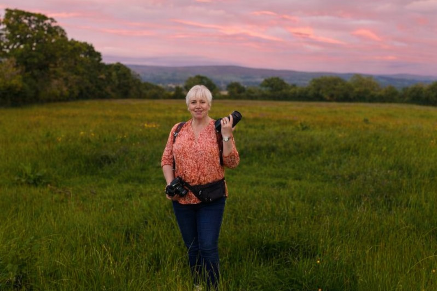 A person stands in a grassy field at sunset with rolling hills and pink sky in the background.