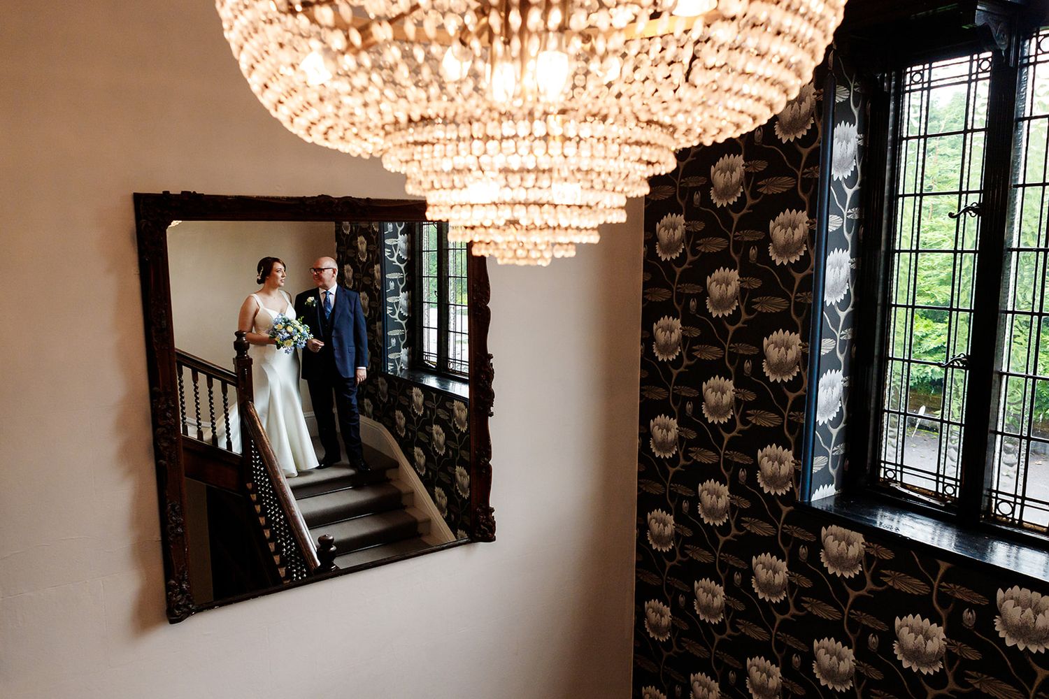 An elegant crystal chandelier hangs above a staircase with black skull-patterned wallpaper and a framed mirror reflection.