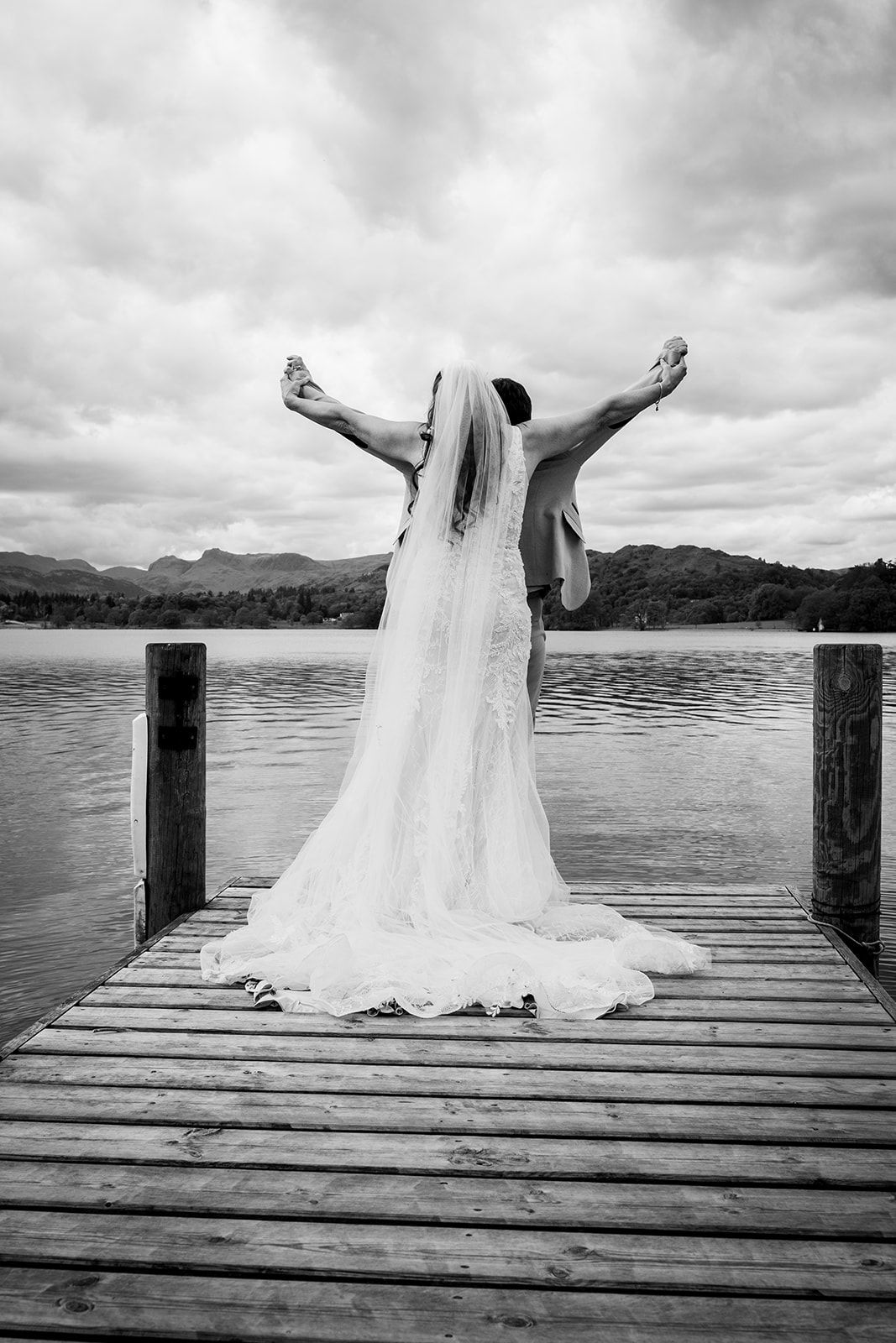Black and white photo of a bride in a flowing gown and veil on a dock, arms outstretched over a scenic lake.