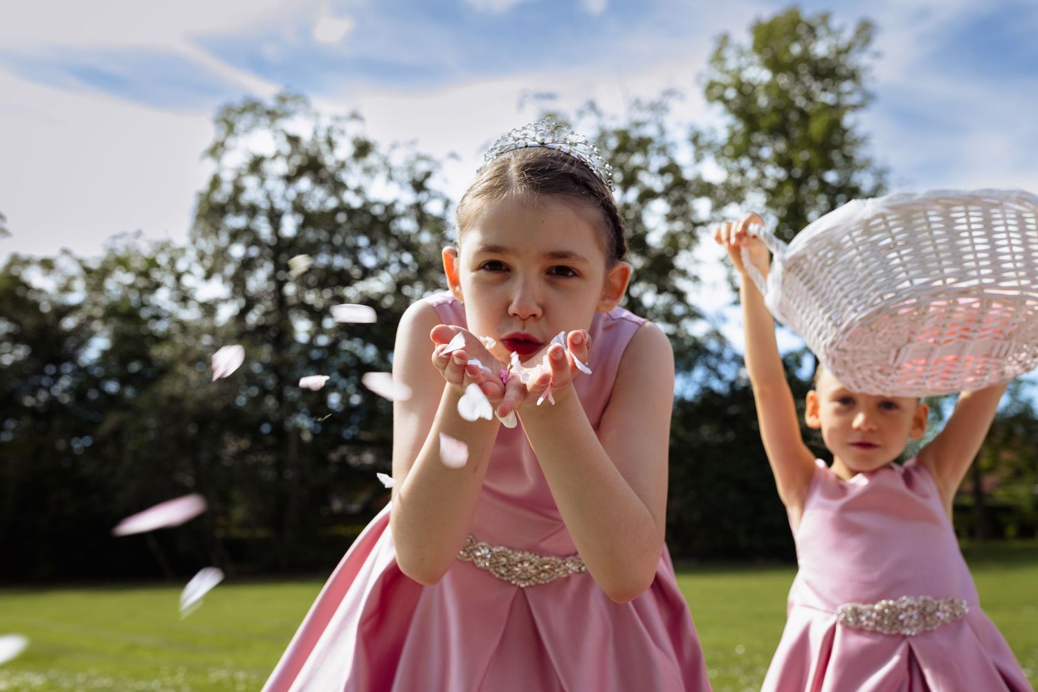 Two young girls in pink dresses and tiaras tossing flower petals outdoors at a wedding ceremony.