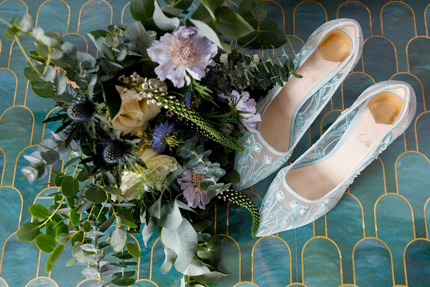 Elegant lace wedding shoes displayed next to a romantic bouquet of mixed flowers on turquoise patterned surface.