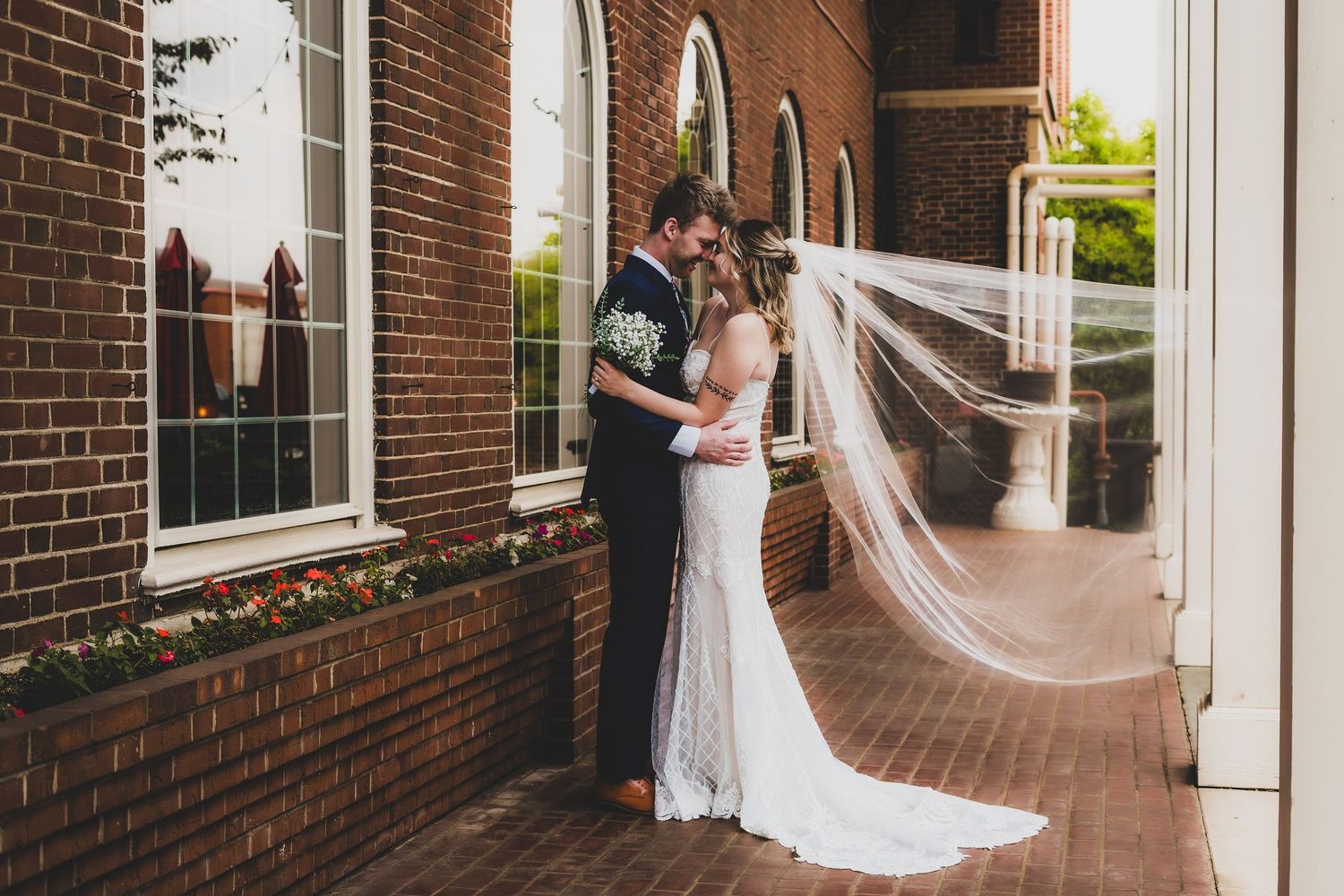 Bride and groom share a romantic embrace outside a brick church, her long veil flowing in the breeze.
