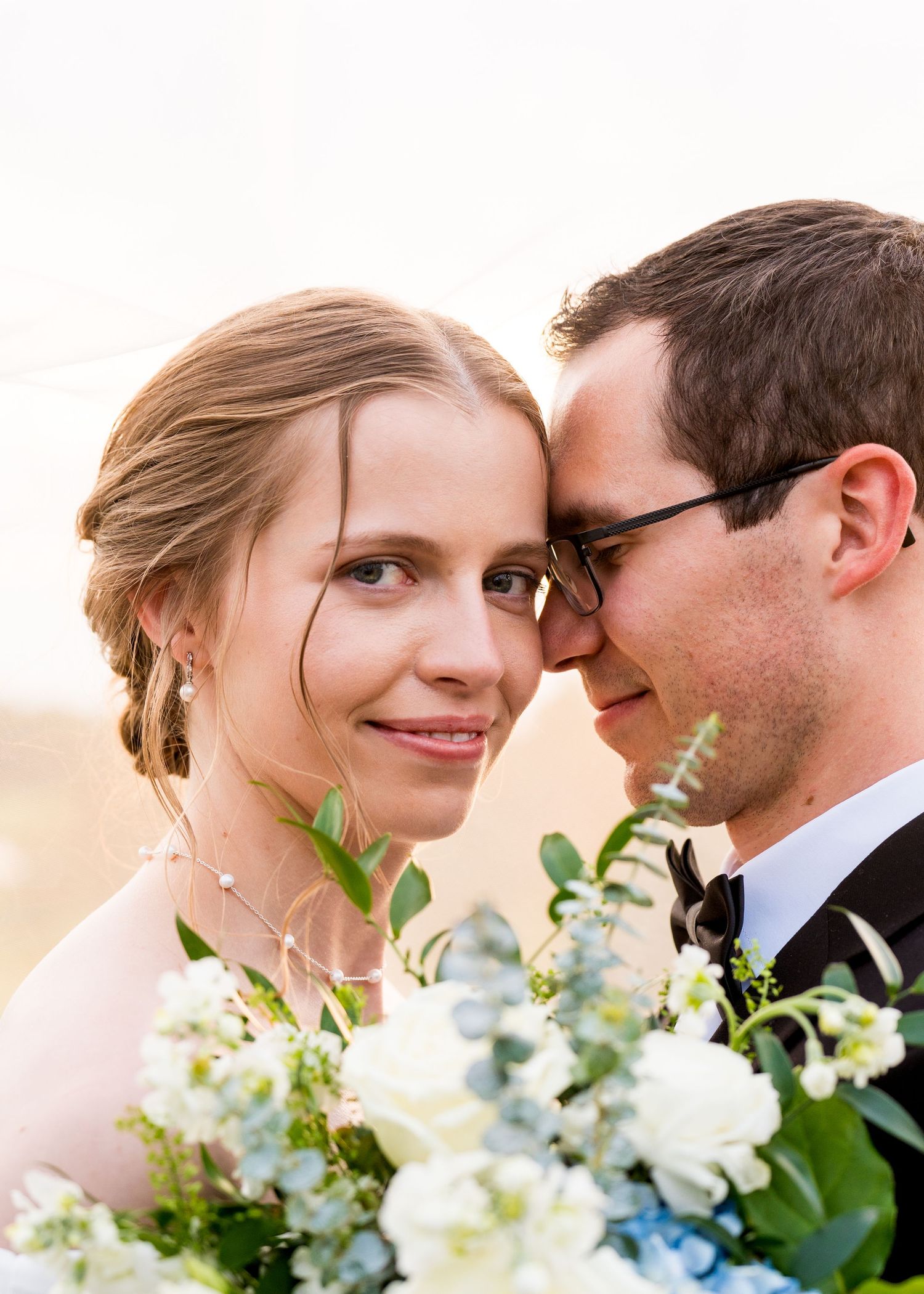 A bride and groom pose cheek-to-cheek, she holding white floral bouquet, wearing elegant wedding attire outdoors.