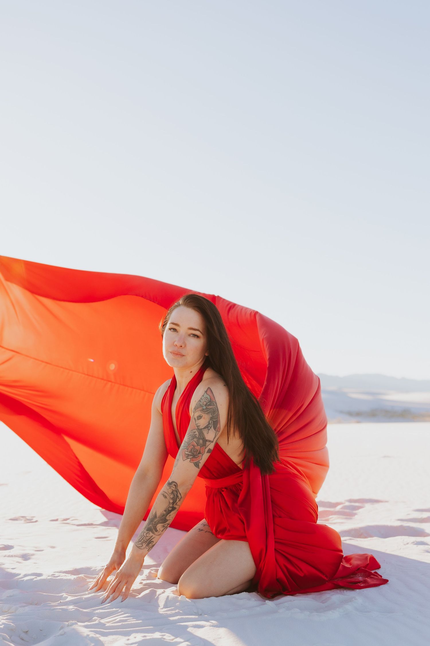A figure in vibrant red fabric creates dynamic movement against white sand and blue sky.