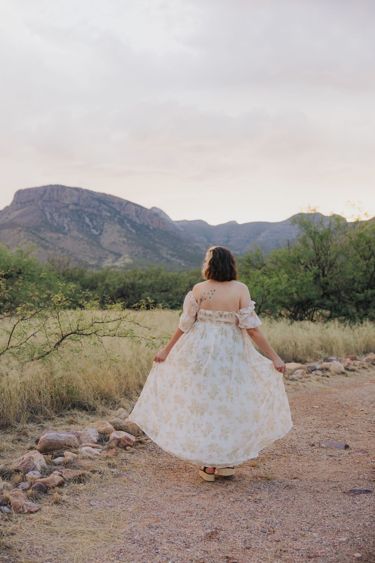 A white floral dress contrasts with the rugged desert landscape and mountain backdrop.