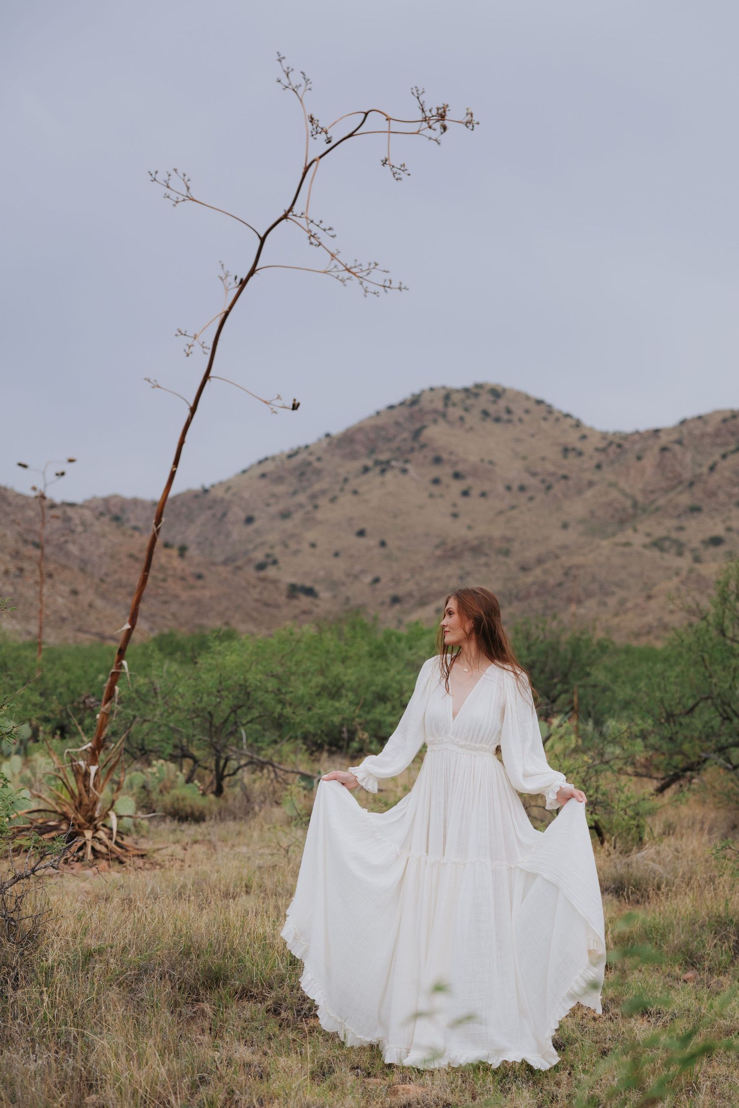 A flowing white dress billows in the desert wind against a backdrop of mountains and desert plants.