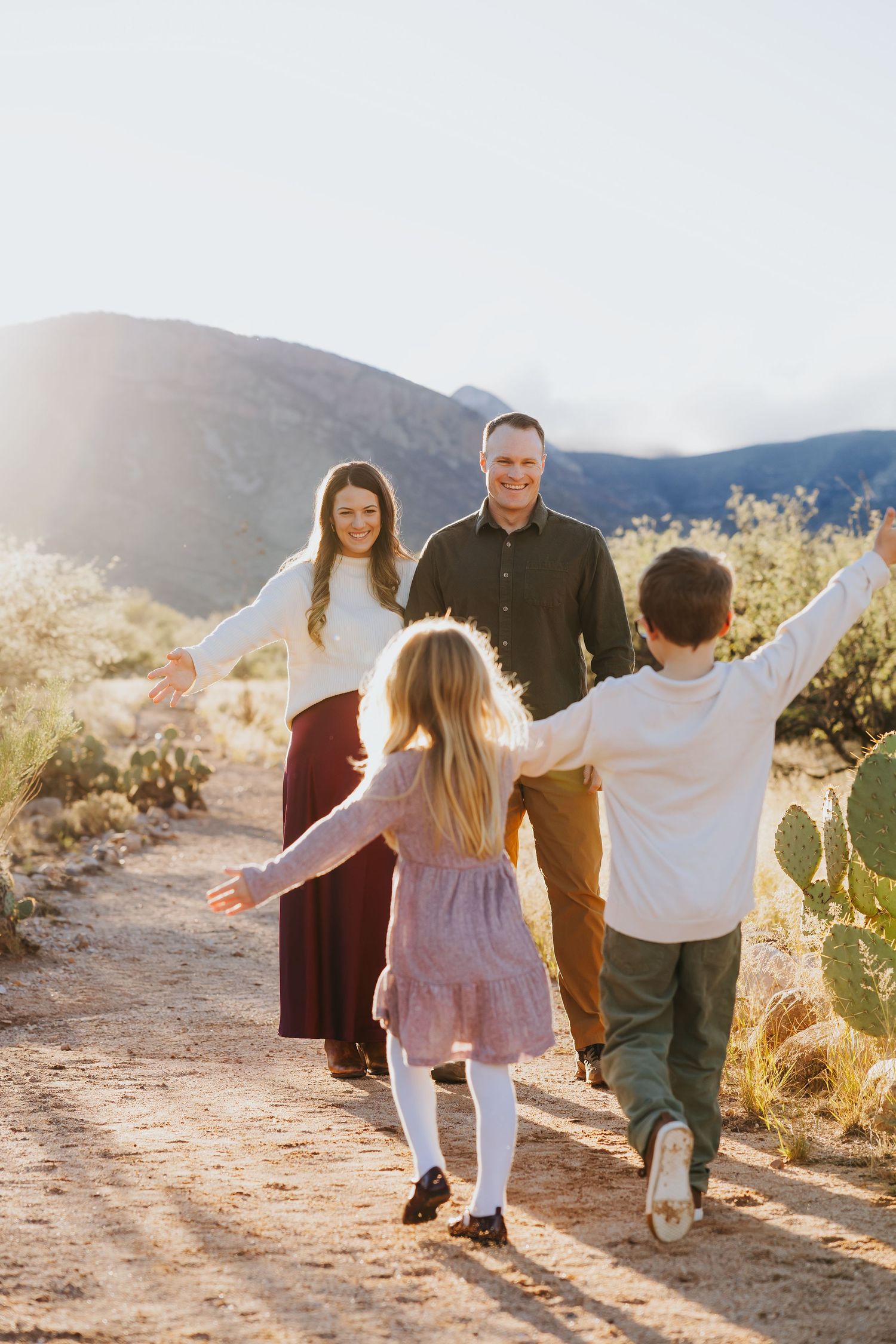 Family walking together on desert trail with arms outstretched during sunset.