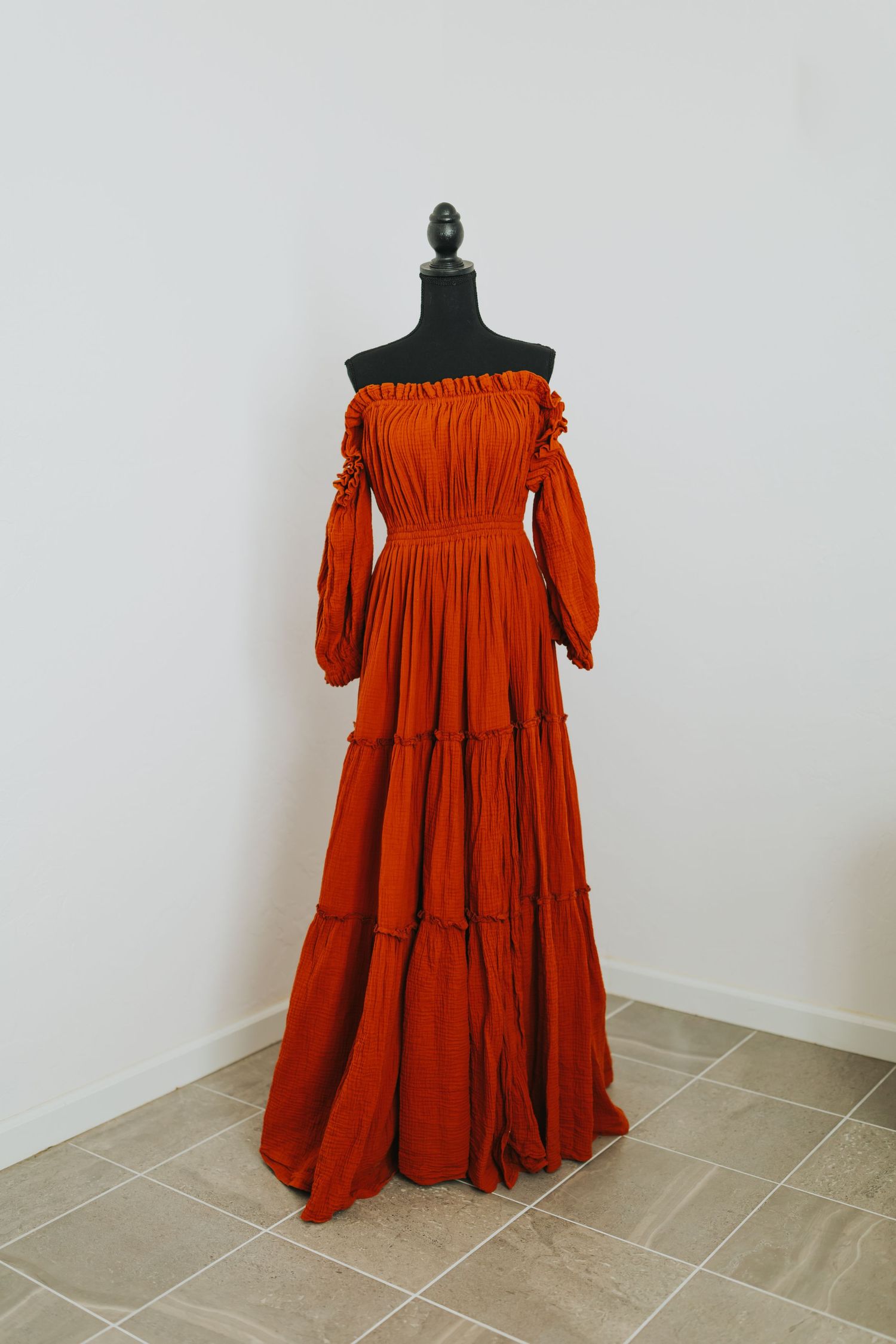 Rust-colored off-shoulder maxi dress with tiered skirt displayed on mannequin against white wall.
