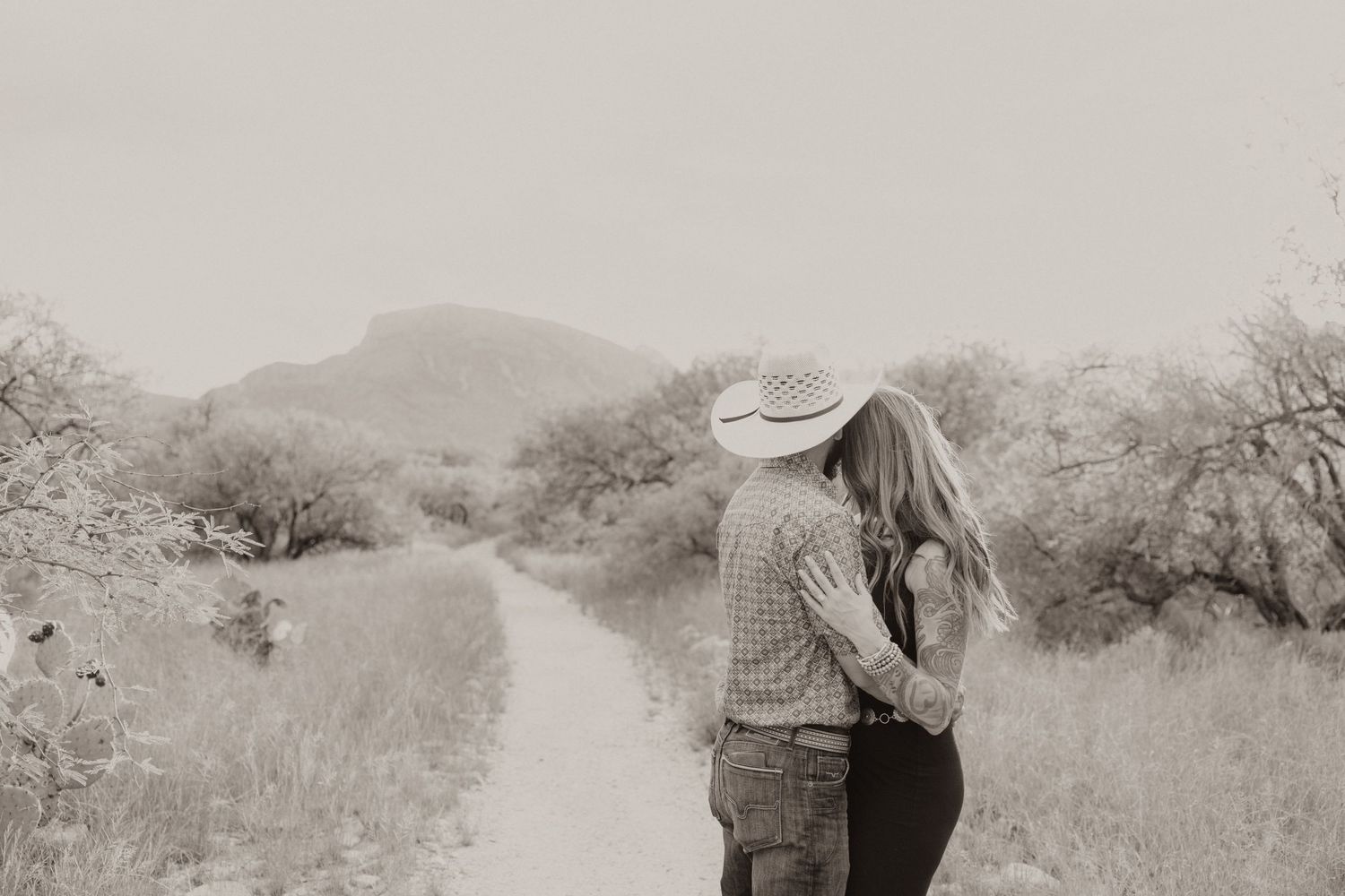 Black and white photo of couple walking together on desert trail with mountain backdrop.