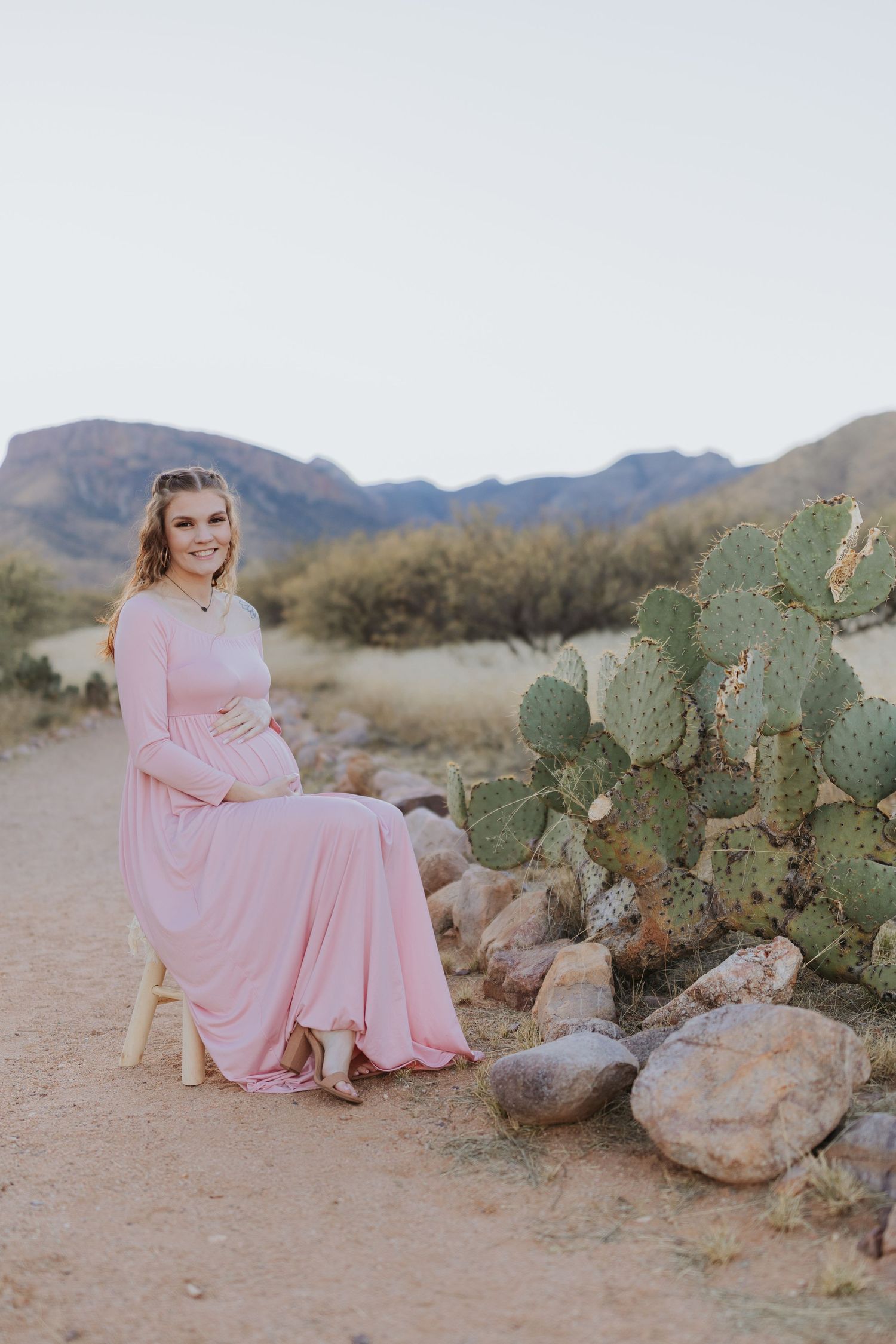 A maternity portrait session with pink dress and desert cacti against mountain backdrop.