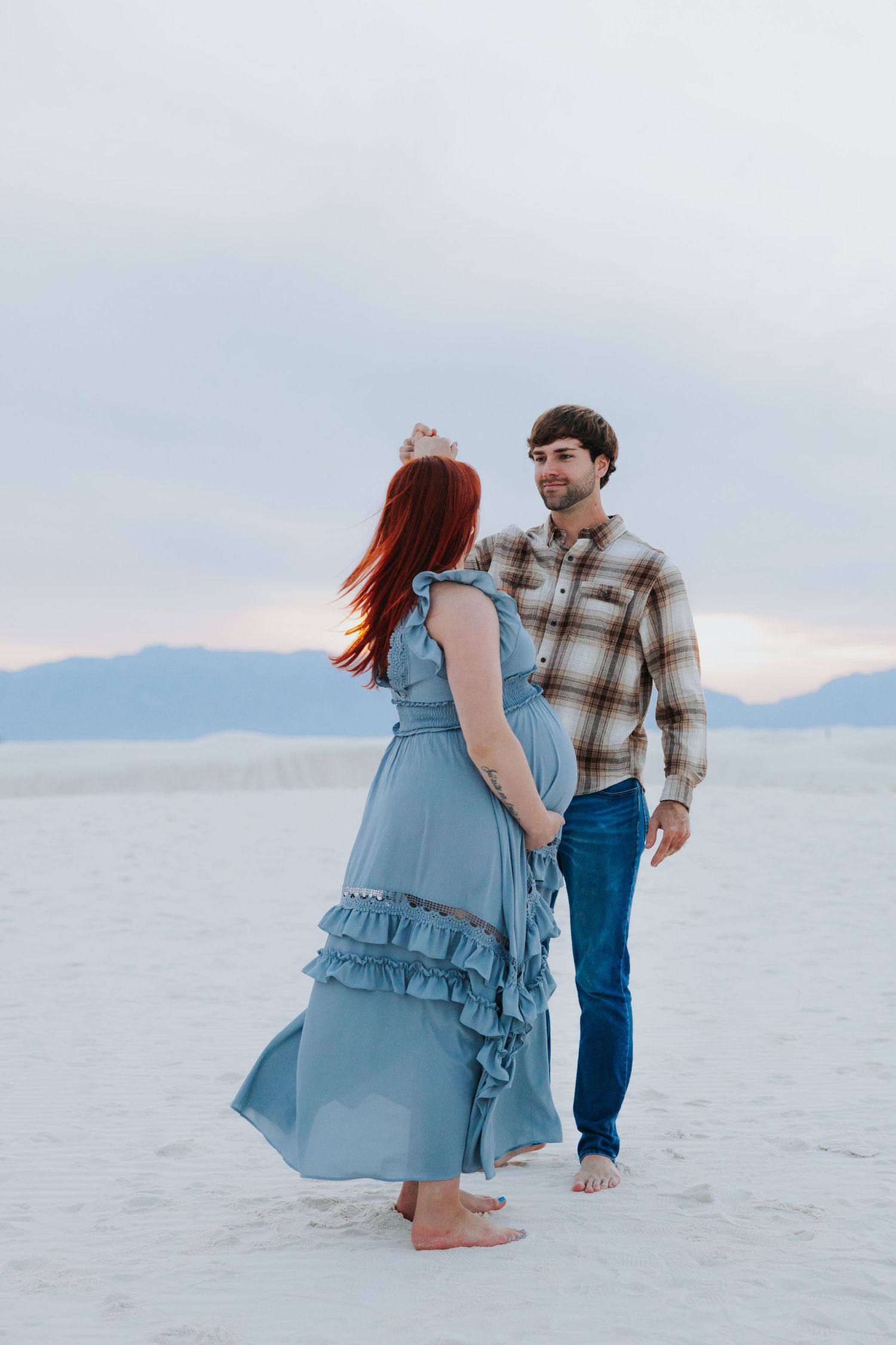 Two people walking barefoot on white sand during sunset with mountains in the distance.