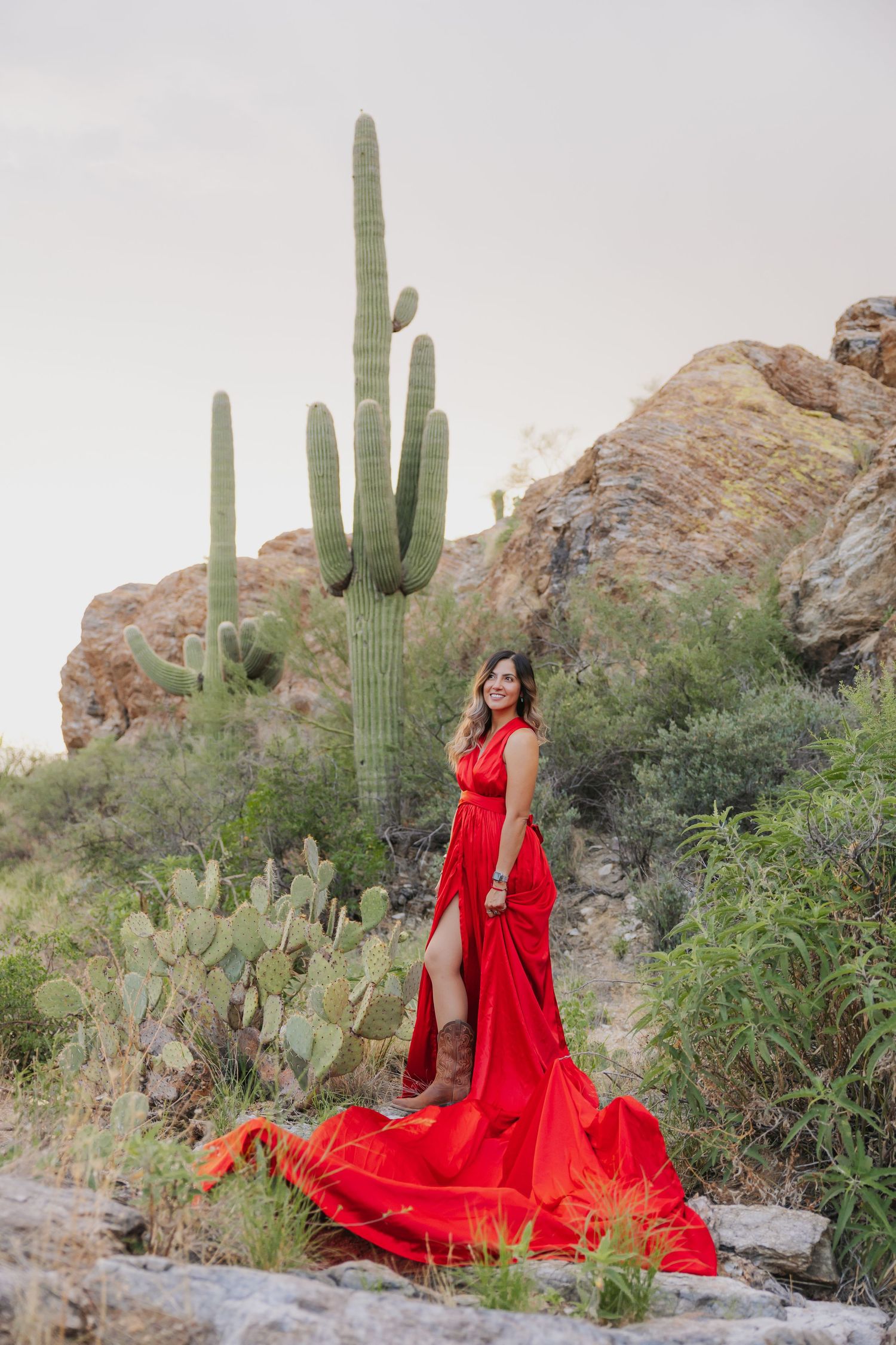 A vibrant red gown adds drama to a desert photoshoot among towering saguaro cacti and rocky outcrops.