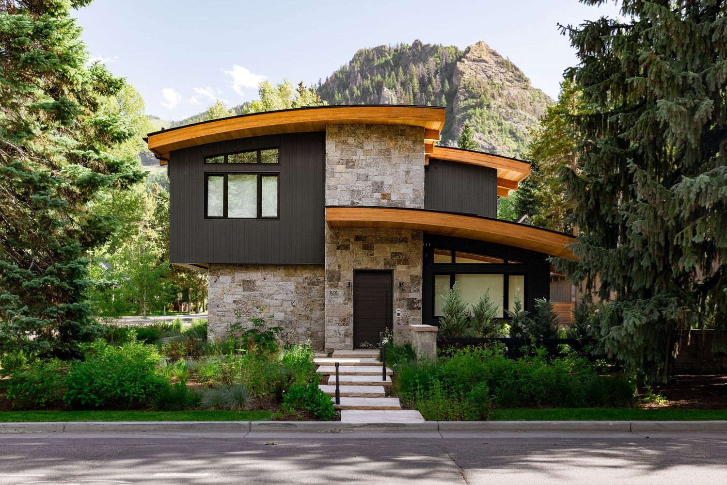 Modern mountain home with curved roof, stone facade, and dark wood paneling, surrounded by pine trees and alpine landscape.