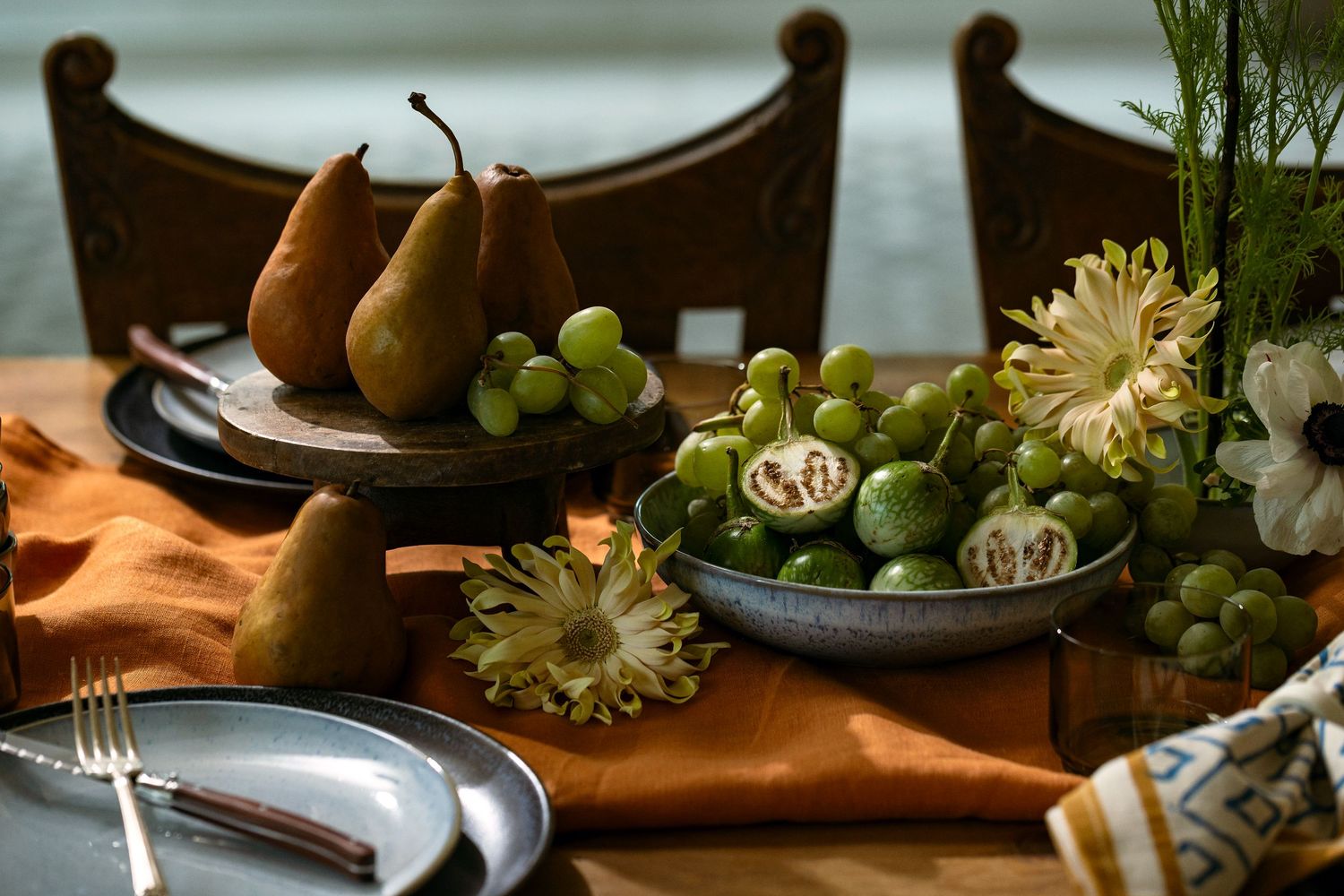 Product photography of ceramic serving plates styled with pears, grapes, and floral accents on a dining table with warm natural light.