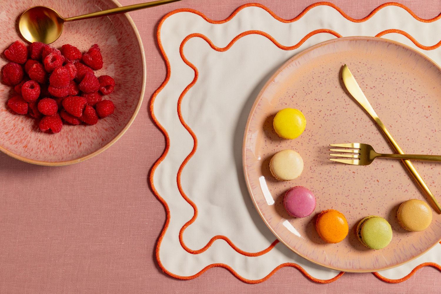 Product photography of a ceramic dessert plate styled with colorful confections, gold flatware, raspberries, and a decorative linen backdrop