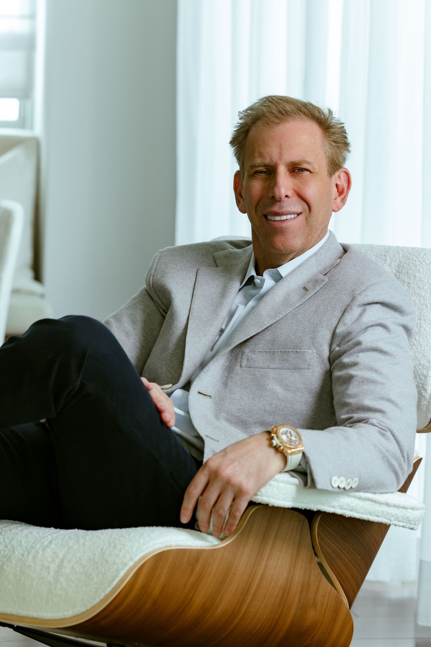 Executive branding photo of Michael Liebowitz, CEO of Douglas Elliman, smiling in a gray blazer while seated in a modern designer chair.
